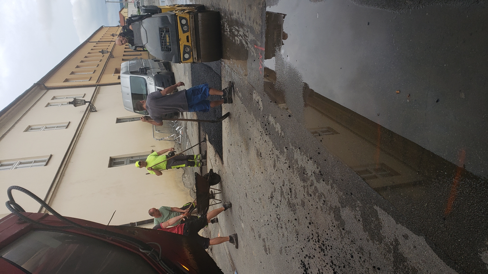 A street paving crew is working on a road in Prešov, Slovakia. The scene shows a dump truck, a asphalt roller, and several workers engaged in the paving process. One worker is operating the asphalt roller, which is compressing fresh asphalt. Another worker is using a shovel to spread asphalt, while a third is pushing a wheelbarrow full of asphalt. The environment appears to be a residential or commercial street, with buildings visible in the background. There is a large puddle of water on the road, reflecting the surrounding buildings and cloudy sky. The weather is overcast, suggesting a possibility of rain.
