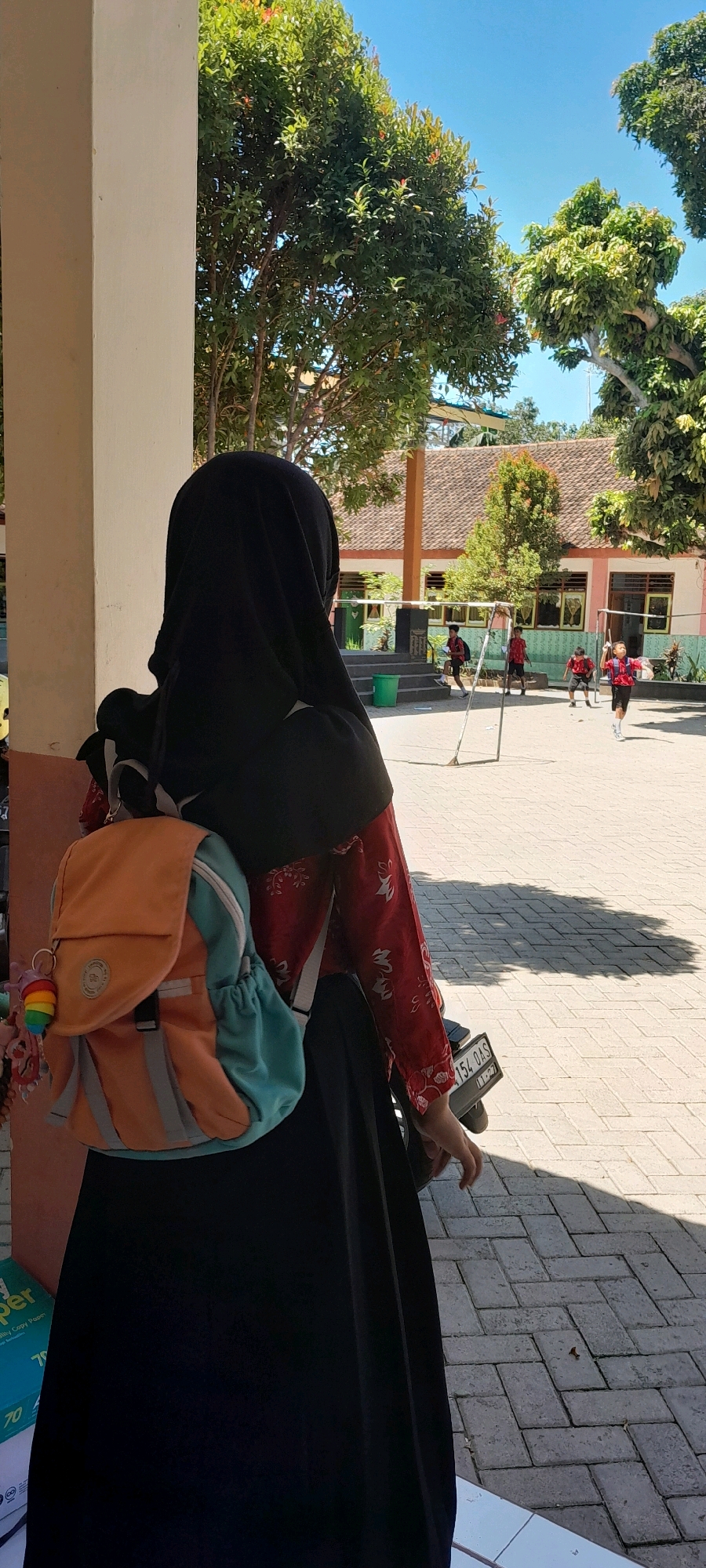 In the city of Lemahbang Dewo, Indonesia, a young person wearing a black hijab and a red patterned shirt is seen from the back, with an orange and teal backpack on. They are standing outdoors on a paved area, possibly a schoolyard, next to a white pillar. In the background, there are several children in red shirts playing with a soccer goal. The scene is bright and sunny, with trees and a building visible.