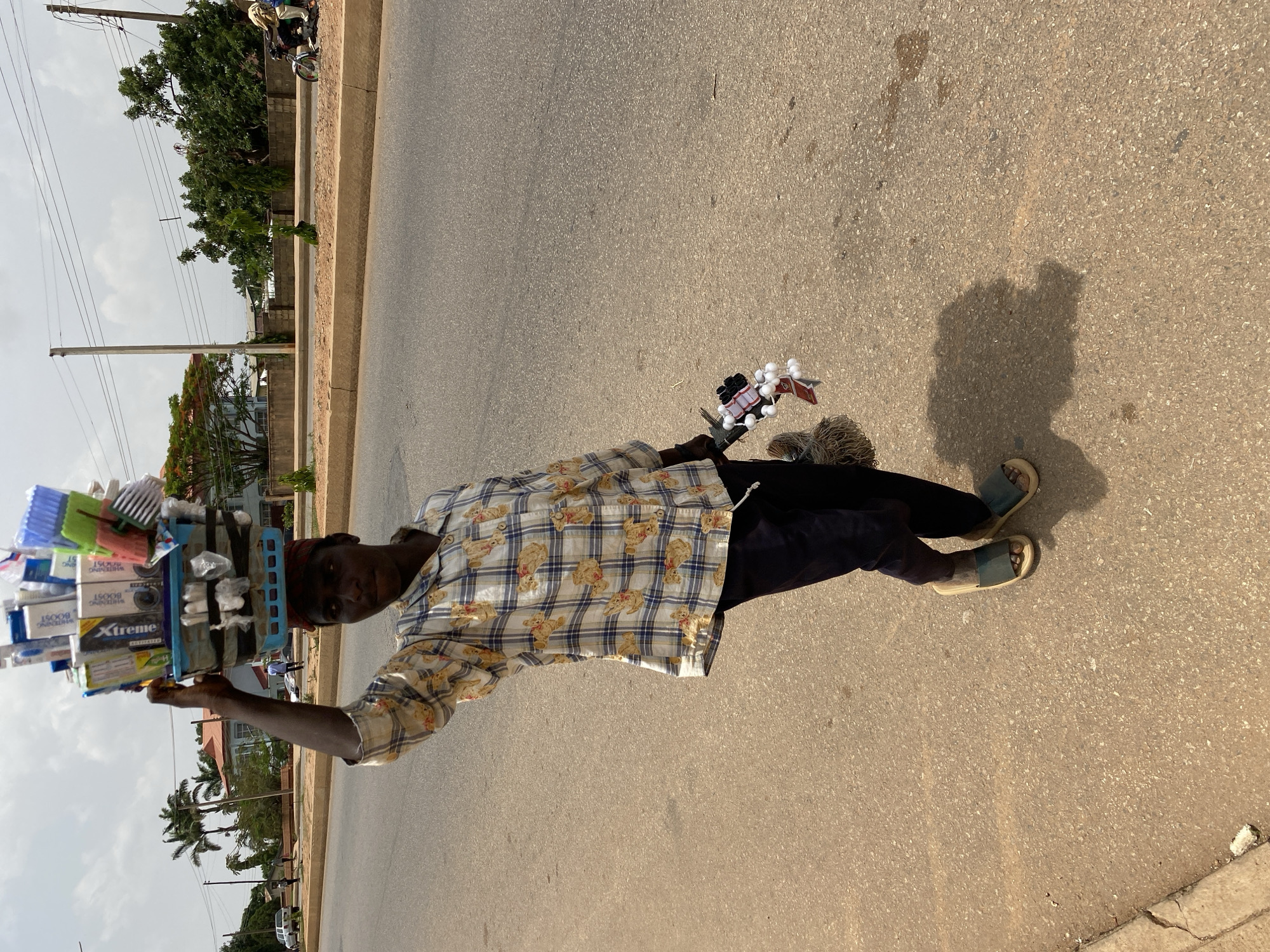 A man stands on a street in Bukuru, Nigeria. He wears a patterned shirt with teddy bear designs, dark pants, and sandals. Atop his head, he carries a blue tray filled with various items, including toothbrushes, toothpaste, and brushes. The street is asphalt with a sidewalk visible on the left side of the image. Buildings and trees line the background. A motorcycle passes in the far background. His shadow falls on the street beside him.
