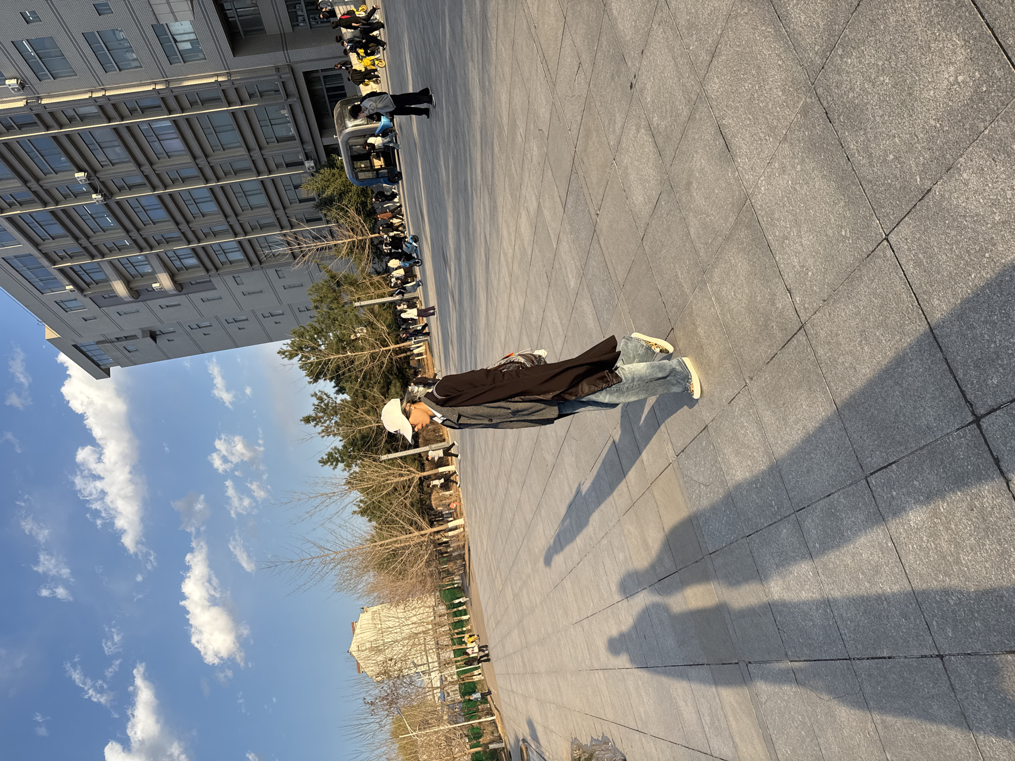 The image shows a person standing on a paved area. They are wearing a white cap, a gray jacket, and jeans. Their shadow is cast on the ground. In the background, there is a building, trees, and a line of vehicles and people. The sky is blue with clouds. The location is Zhongtun, China.