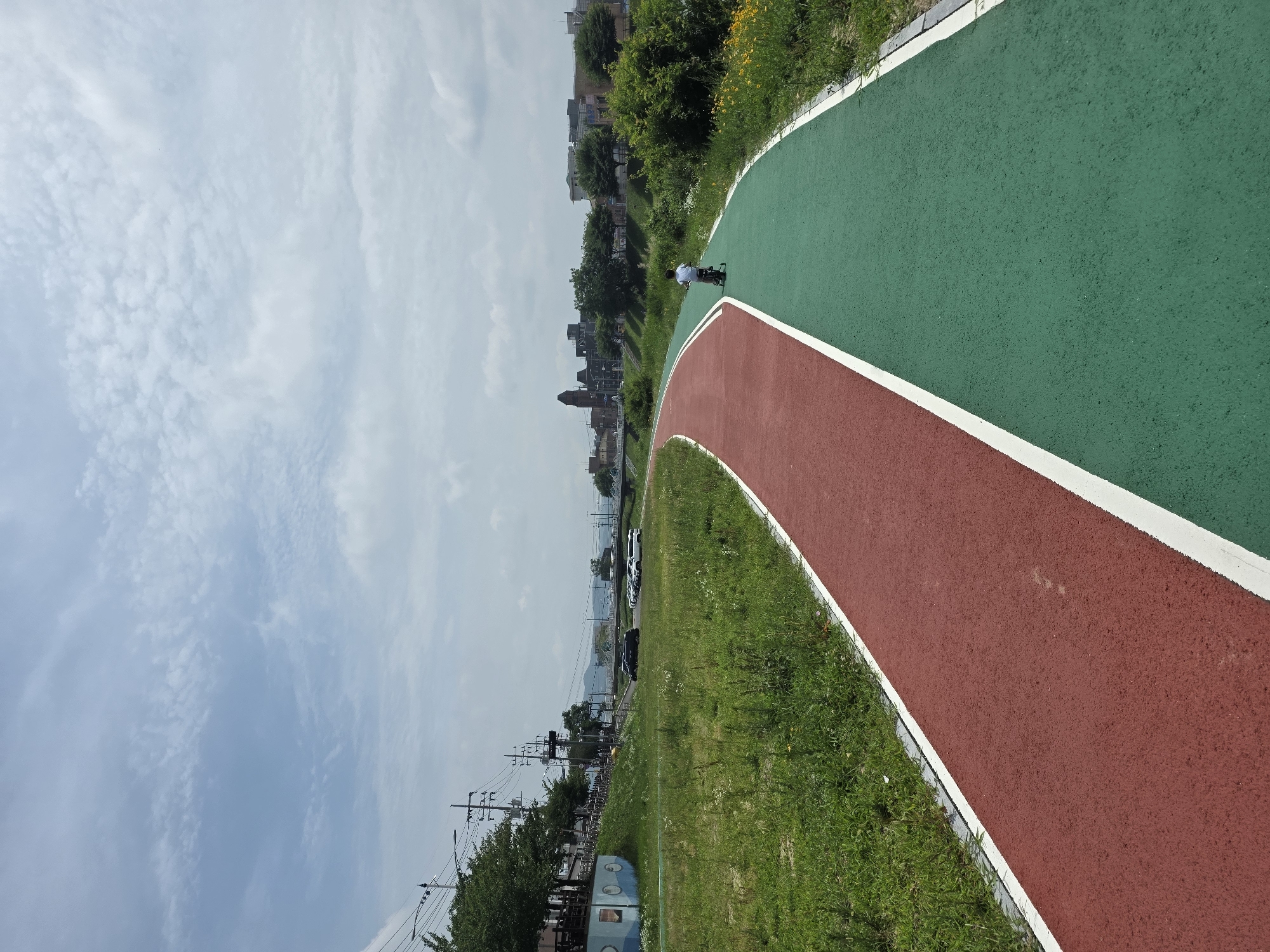 A young boy rides a tricycle on a paved bike path that is divided into a red lane and a green lane. The path curves to the right and runs alongside a grassy field. In the distance, there are buildings and trees, and a bridge crosses a body of water. The sky is cloudy. The image appears to be taken in Anseong-si, South Korea, possibly in a park or recreational area along a river.