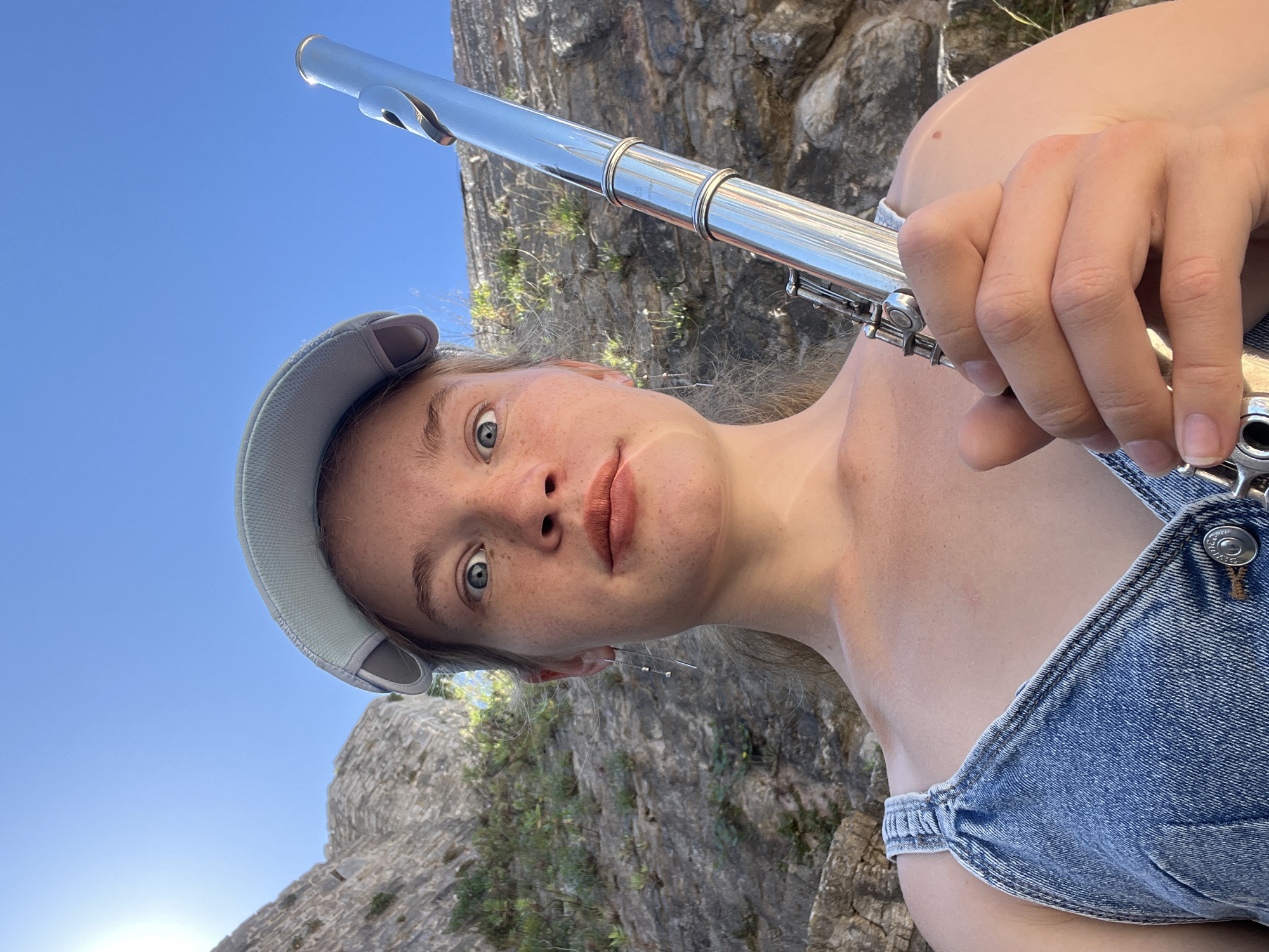 A young woman wearing a blue denim overall dress and a light blue baseball cap holds a silver flute in her right hand. Her eyes are wide and directed at the camera, and her lips are slightly parted. She has freckles across her nose and cheeks, and is wearing a long, dangling earring in her left ear. Behind her, a rugged, rocky hillside with sparse vegetation rises against a bright blue sky. The scene appears to be outdoors, possibly in a mountainous or coastal area. The location is identified as Budva, Montenegro.