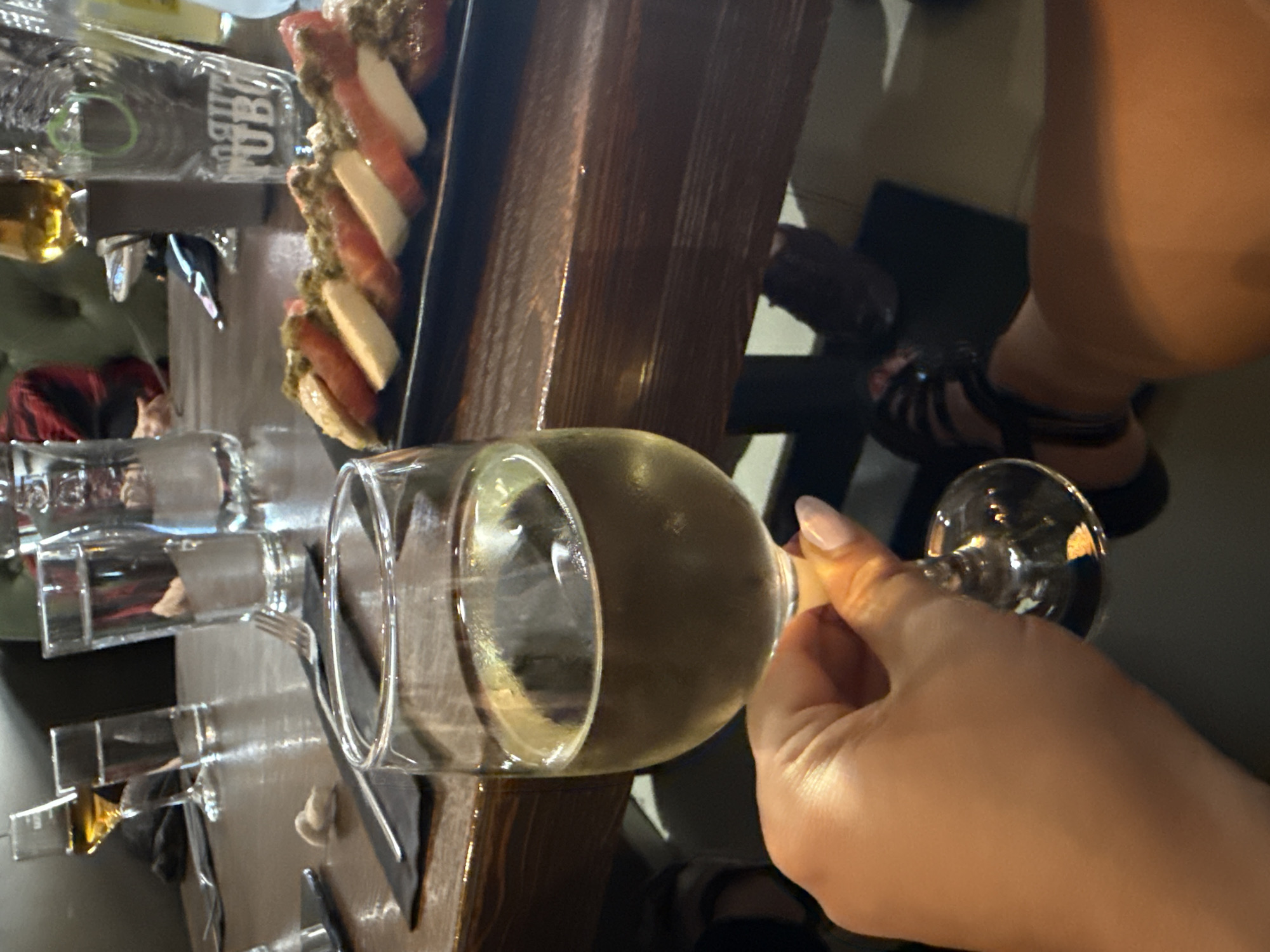 An indoor scene depicts a person's right hand holding a stemmed glass of white wine in the foreground. The hand, with manicured nails painted in a light neutral polish, grasps the stem of the glass, which is partially filled with a pale yellow liquid. The person's right leg, wearing a black strappy sandal, is visible in the lower right portion of the image.
The setting is a dining establishment in the city of Unknown, Unknown, featuring a dark brown wooden table. On the table, a black rectangular plate holds a dish composed of alternating slices of red tomatoes and white mozzarella cheese, generously topped with a green, textured sauce, resembling a Caprese salad. To the right of the plate, a tall, clear glass with a "TUBORG" logo is visible. Further back, two other tall, clear glasses, one appearing full of water, are positioned on the table. A stemmed glass containing an amber liquid (possibly an alcoholic beverage) is also visible in the background. A silver fork rests on a black napkin or placemat on the table. Another small, light-colored crumpled object is present on the table surface.
The background shows dark-colored upholstered seating and partial views of other diners, including a person wearing a red and black plaid shirt. The lighting is dim, casting reflections on the glass surfaces and the glossy wooden table.