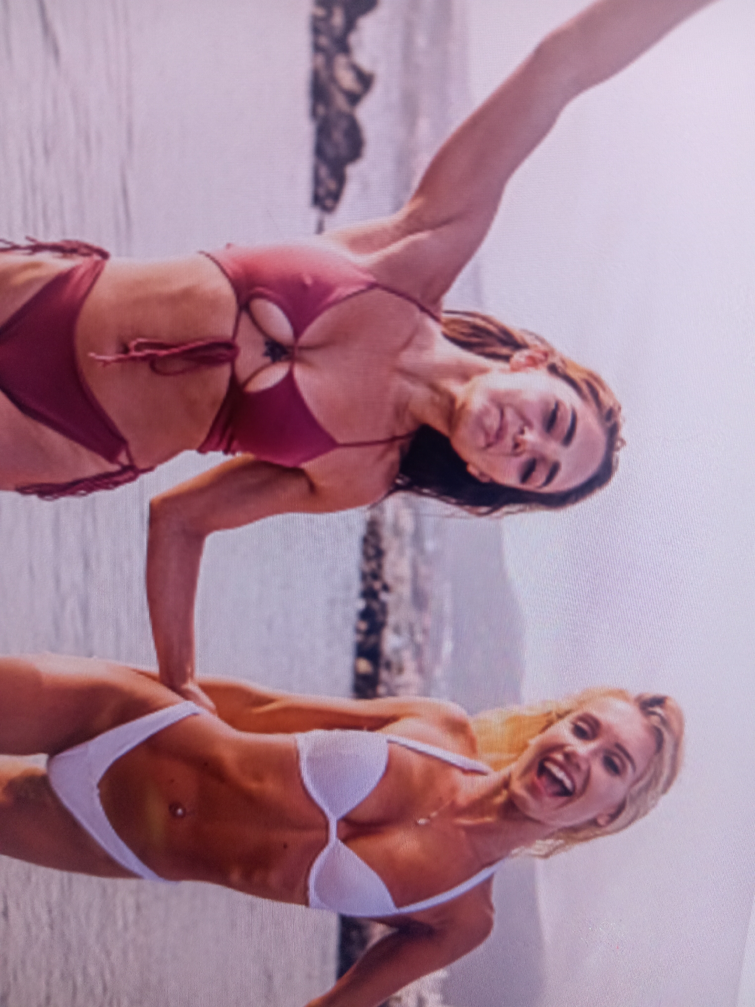 Two women are in the water at a beach. The woman in the foreground is wearing a white bikini. The woman in the background is wearing a maroon bikini. They are both smiling and appear to be having fun. The beach is rocky and there are some palm trees in the background. The water is clear and blue.