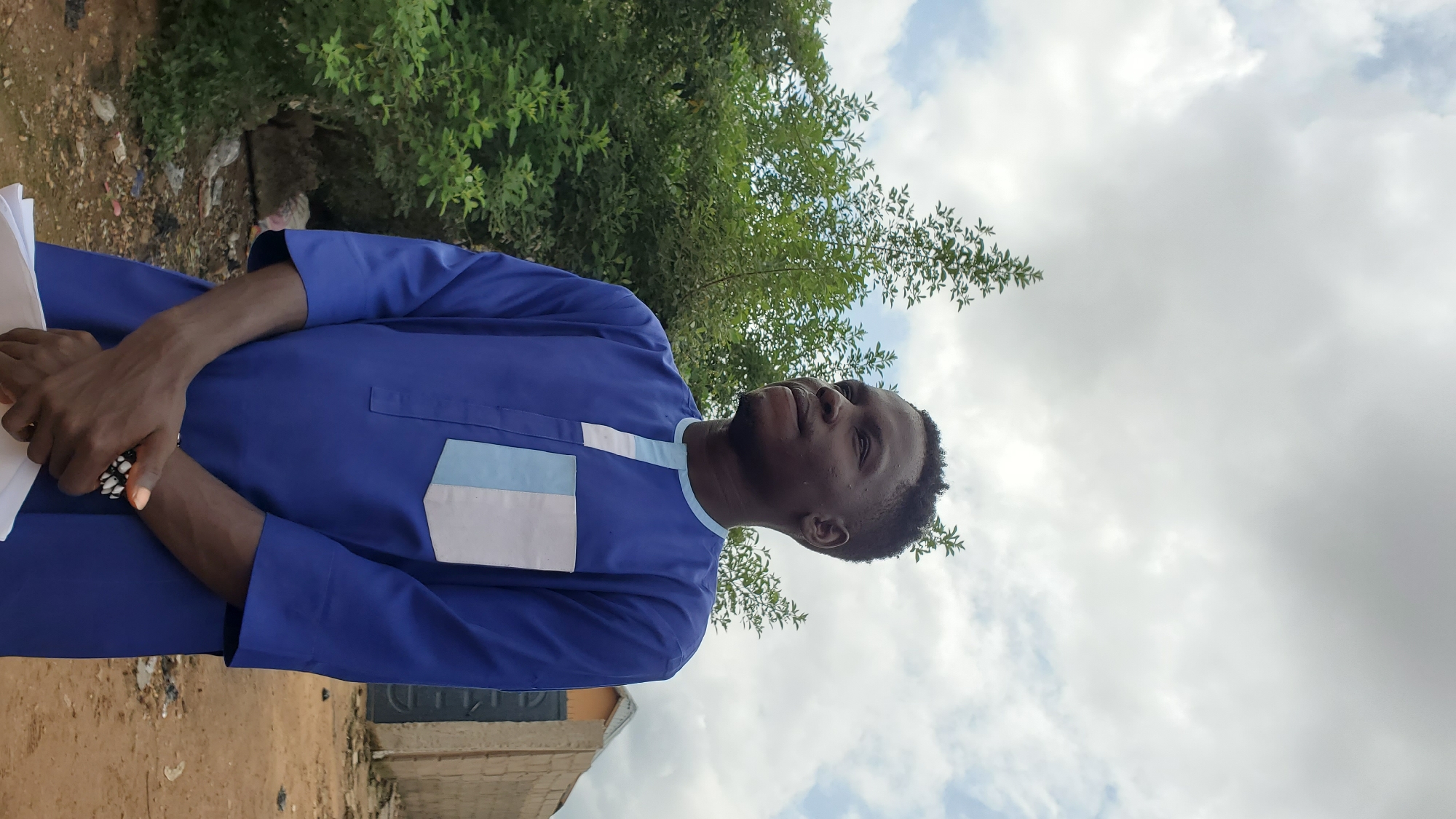 In Jalingo, Nigeria, a young Black man stands outdoors in a blue tunic with white and light blue accents on the chest. His hands are clasped in front of him, and he is wearing a bracelet on his left wrist. He is looking to his right, with lush green trees and bushes behind him. A light brown building and a gray gate are visible in the background, with a cloudy sky overhead. The ground is sandy and uneven, with some scattered debris.