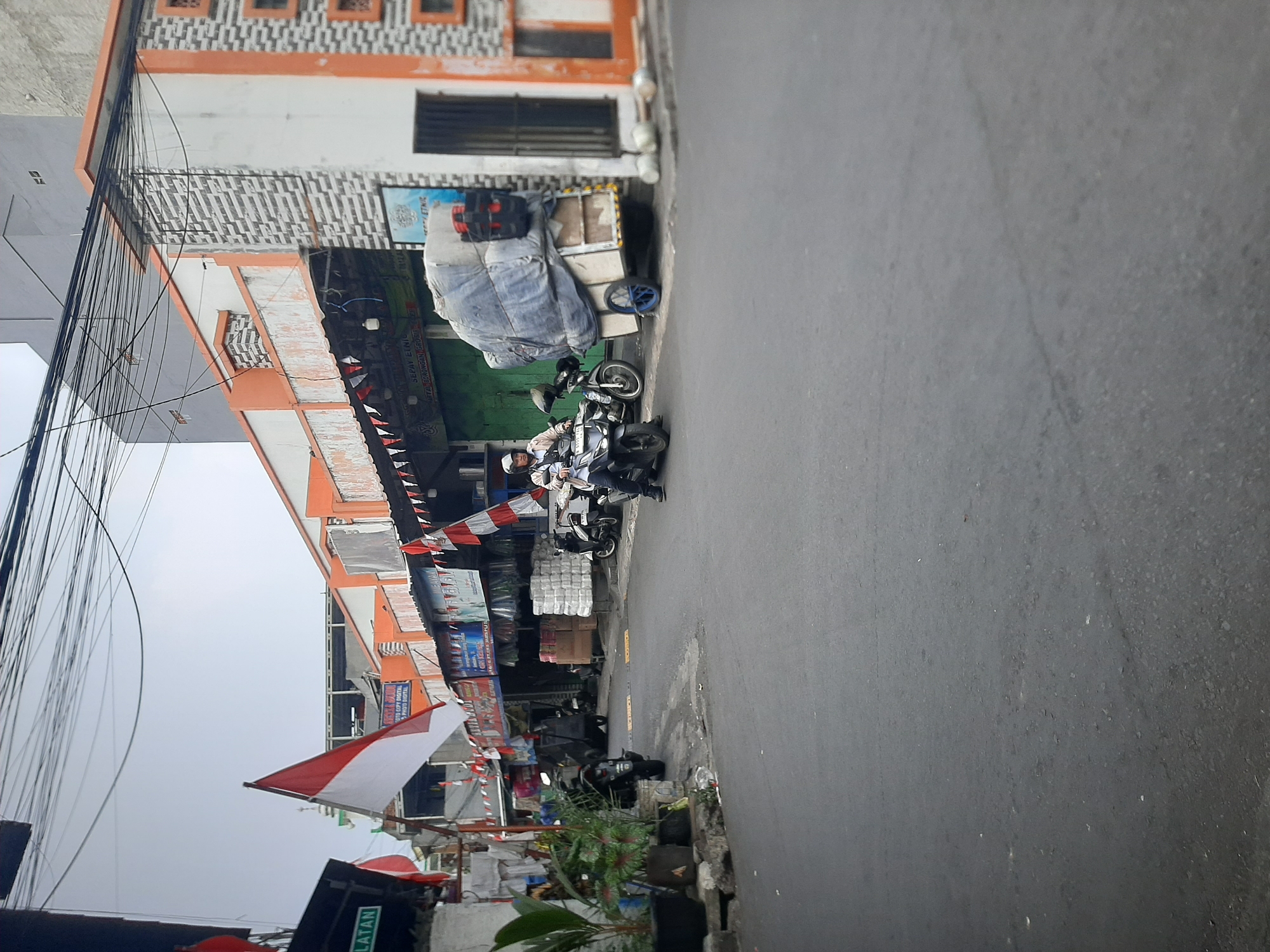 The image depicts a street scene in Central Jakarta, Indonesia, likely on a cloudy day. The street is paved and appears to be in an urban setting with buildings lining one side.
On the left side of the street, there are buildings with awnings and some plants in pots. An Indonesian flag is prominently displayed, along with smaller red and white pennant flags. There are several motorcycles parked, and one is in motion.
On the right side of the street, there are more buildings, some with orange and gray patterned facades. A cart loaded with bulky items covered in a blue tarp is parked next to a building. There are also stacks of what appear to be white goods or supplies. Another motorcycle is visible, and a person wearing a helmet is riding it. The road surface shows some wear and tear. Overhead, a dense network of electrical wires is visible against the sky.