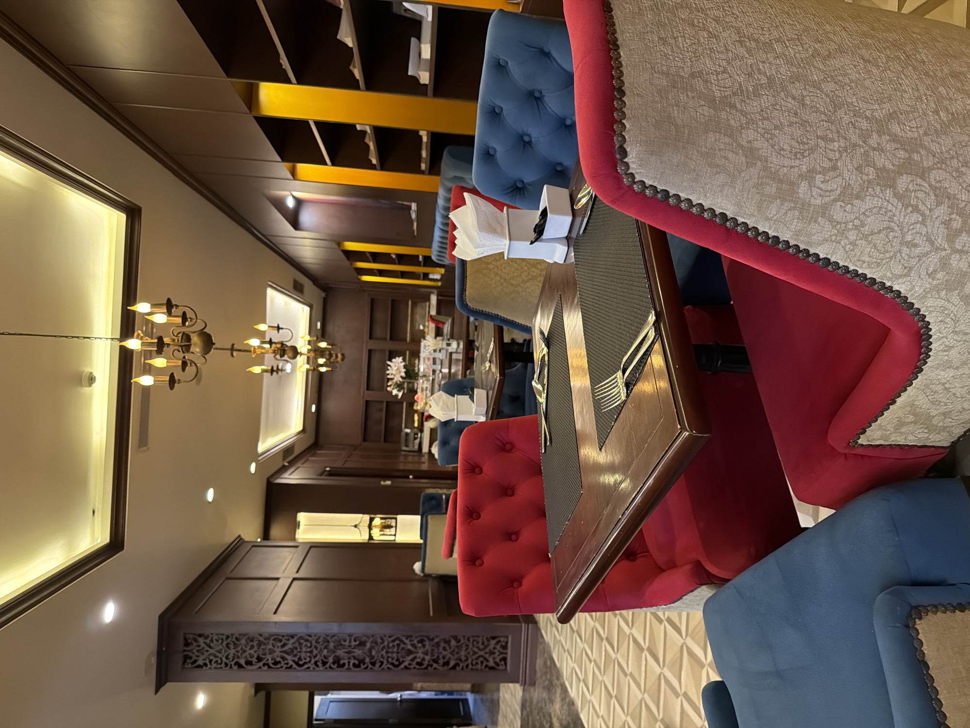 The image shows an indoor dining area, likely a restaurant or hotel lobby, with a formal setting. Several tables are arranged with plush seating in vibrant red and deep blue upholstered chairs, some featuring decorative nailhead trim and patterned fabric. Each table is set with dark placemats, silverware, and napkins. The room is dimly lit, with warm ambient lighting emanating from chandeliers and recessed ceiling lights. Dark wood paneling lines the walls, contrasted by ornate carved wooden pillars and gold-colored shelving units. The floor is tiled in a geometric pattern. In the background, a doorway leads to another area, and on a table, a floral arrangement is visible. The overall impression is one of elegance and comfort, suggesting a place for fine dining or relaxation. The specific location in Thuận Hóa Ward, Vietnam, is not visually discernible from the image content.