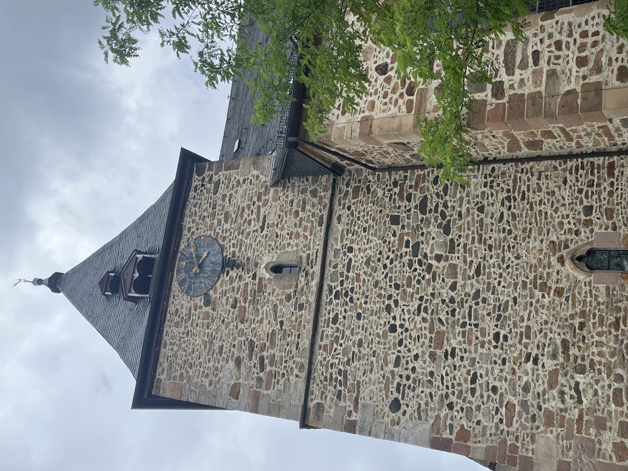 This is a low-angle, vertical shot of the stone facade of a church tower. The rough-hewn stone blocks are a mix of brown, grey, and tan, with white mortar holding them together. The wall is punctuated by small, arched windows and a prominent round clock face with Roman numerals. The clock hands indicate the time is approximately 2:10. The tower has a steeply pitched, dark-tiled roof that transitions to a darker, slate-like material at the peak, topped with a weather vane.
Green foliage from a tree hangs into the frame from the top right, partially obscuring the roofline and a section of the stone wall. The sky is overcast and a pale grey. The stone construction and the Gothic-style arched windows suggest an older European architectural style. Without further context, the specific location within Neustadt, Germany cannot be determined, but the style of architecture is typical of historical buildings in the region. There is no visible text in the image. The overall mood is one of quiet solemnity, with the overcast sky contributing to a muted atmosphere. The image captures a detail of the church's exterior, focusing on its weathered stone and architectural features.