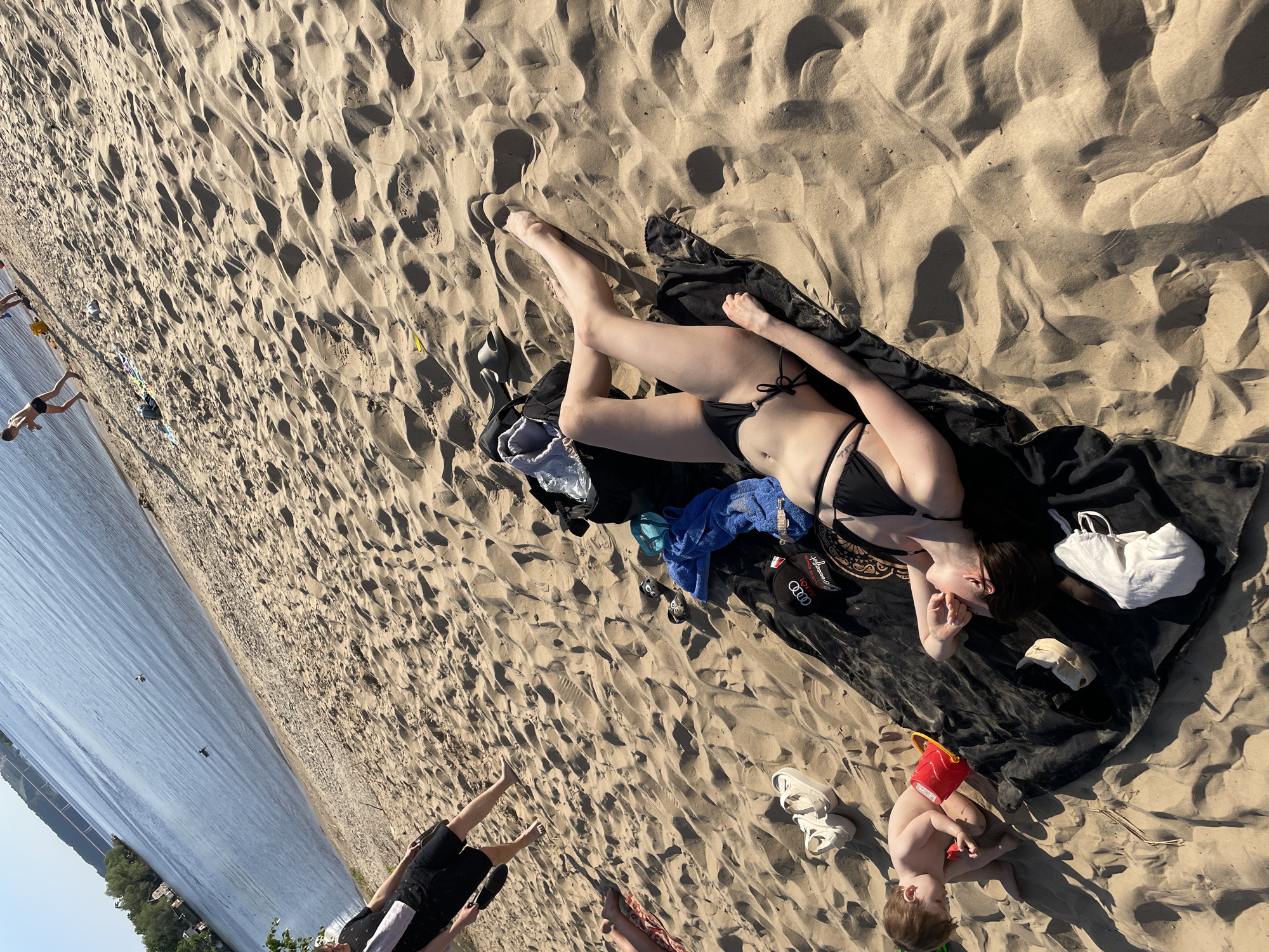 A woman in a black bikini lies on a dark-colored blanket on a sandy beach. A young child in a red swimming suit sits near the blanket, playing with a red bucket. Other items scattered around the blanket include a white bag, a white baseball cap, shoes, a blue towel, and a black hat. In the background, people are visible in the water. The location is Матюшино, Russia, based on the landscape.