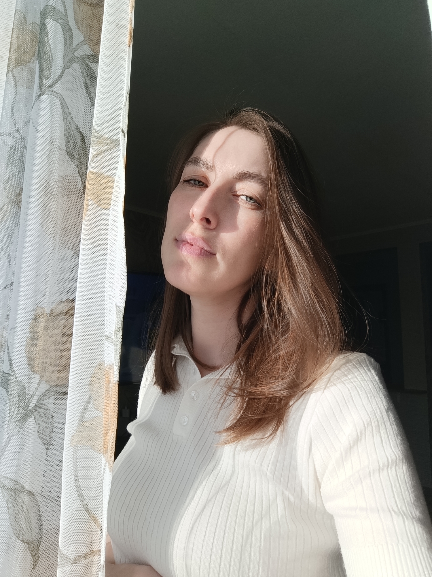 A young woman with shoulder-length brown hair stands in front of a window, partially obscured by a white curtain patterned with floral designs. She is wearing a white, ribbed polo shirt. Her face is illuminated by sunlight, casting shadows across her face. The background behind her appears to be an interior, possibly a room with dark walls. The photograph does not allow for confirmation or denial of the stated location.