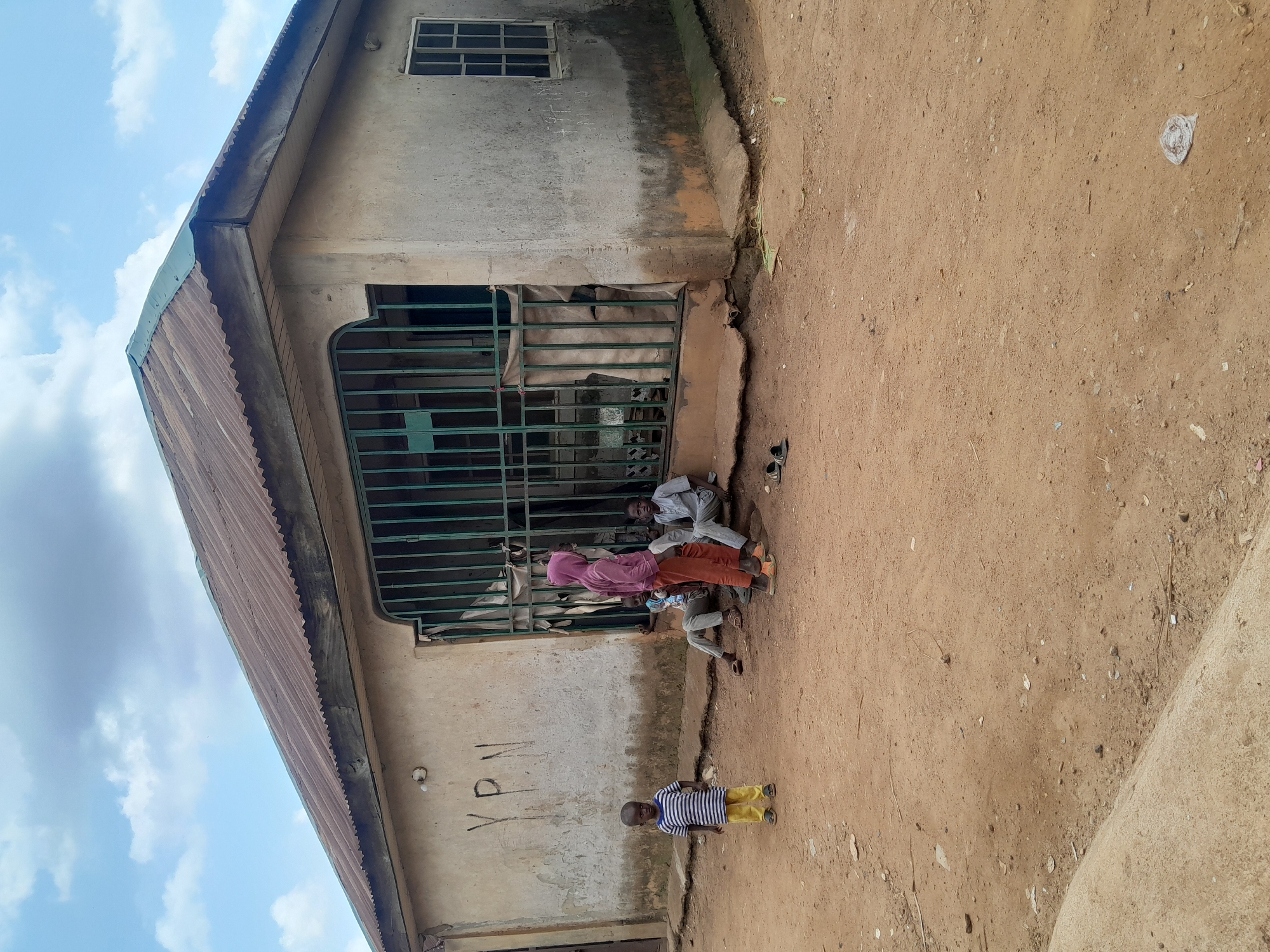 In Madalla, Nigeria, a light-colored, single-story building with a corrugated metal roof is visible. The building has a concrete foundation and a stucco exterior. A green metal gate covers an opening in the front of the building. Several children are gathered outside the building. One child, wearing a striped shirt and yellow pants, stands to the left of the entrance. Two other children, one in a pink hoodie and orange pants and another in a light blue shirt and grey pants, are sitting near the entrance. A fourth child, also in a light blue shirt, sits further inside the entrance. The ground in front of the building is dirt, with scattered debris. A window with a metal grate is visible on the right side of the building. The sky above is partly cloudy.