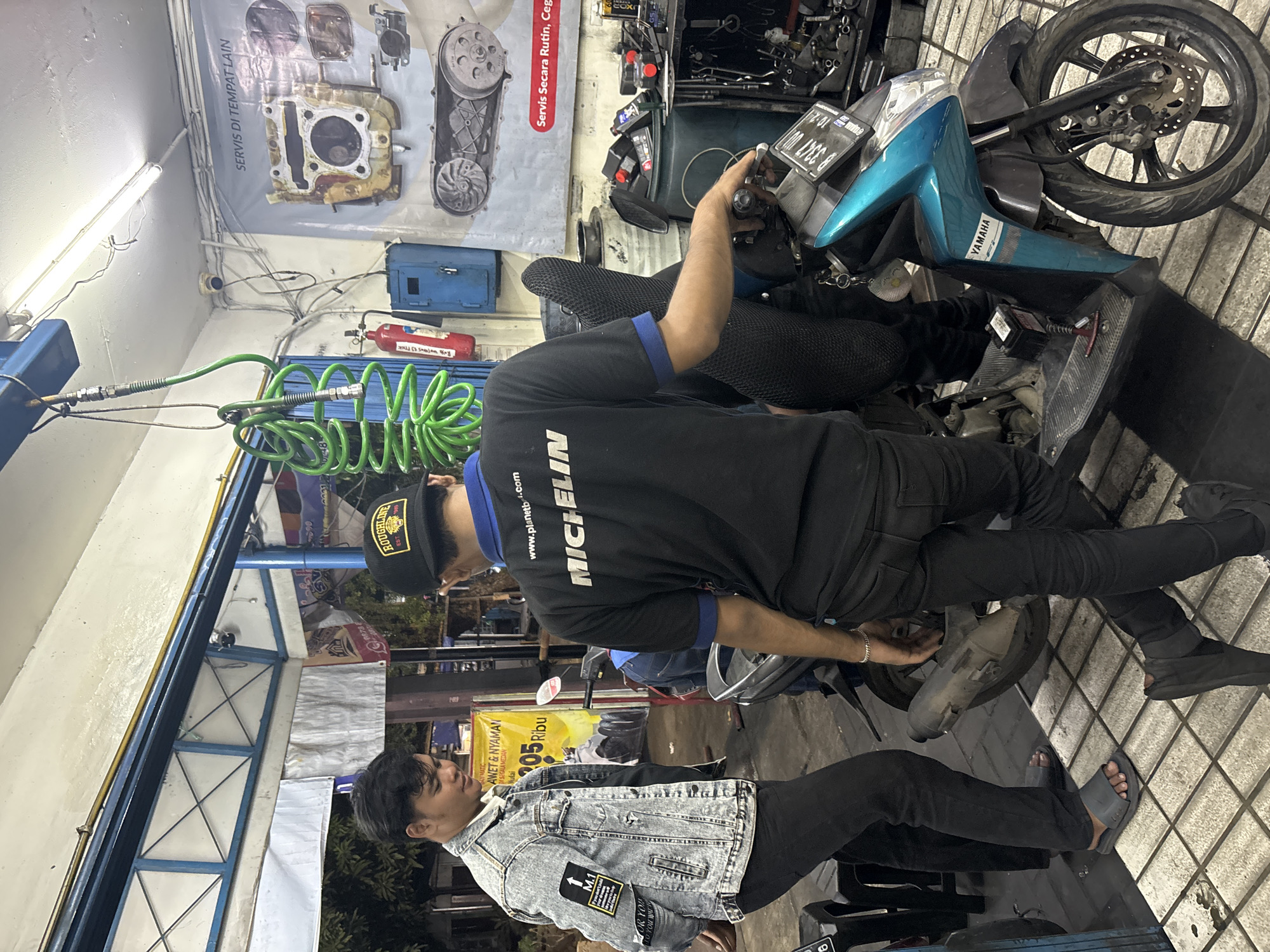 In North Jakarta, Indonesia, at a motorcycle repair shop, two young men are seen interacting with a teal Yamaha scooter. The man on the left, wearing a denim jacket with patches and black pants, stands with his hands clasped behind his back, observing. The man on the right, dressed in a black shirt with "MICHELIN" printed on it and black pants, is in the process of working on the scooter, his right hand on the handlebar. The scooter is parked on a tiled floor, with various tools and equipment visible in the background, including a red fire extinguisher, a blue cabinet, and a poster displaying engine parts. An air hose hangs from the ceiling above them, and overhead fluorescent lights illuminate the workspace. The overall environment suggests an active service area within a city setting.