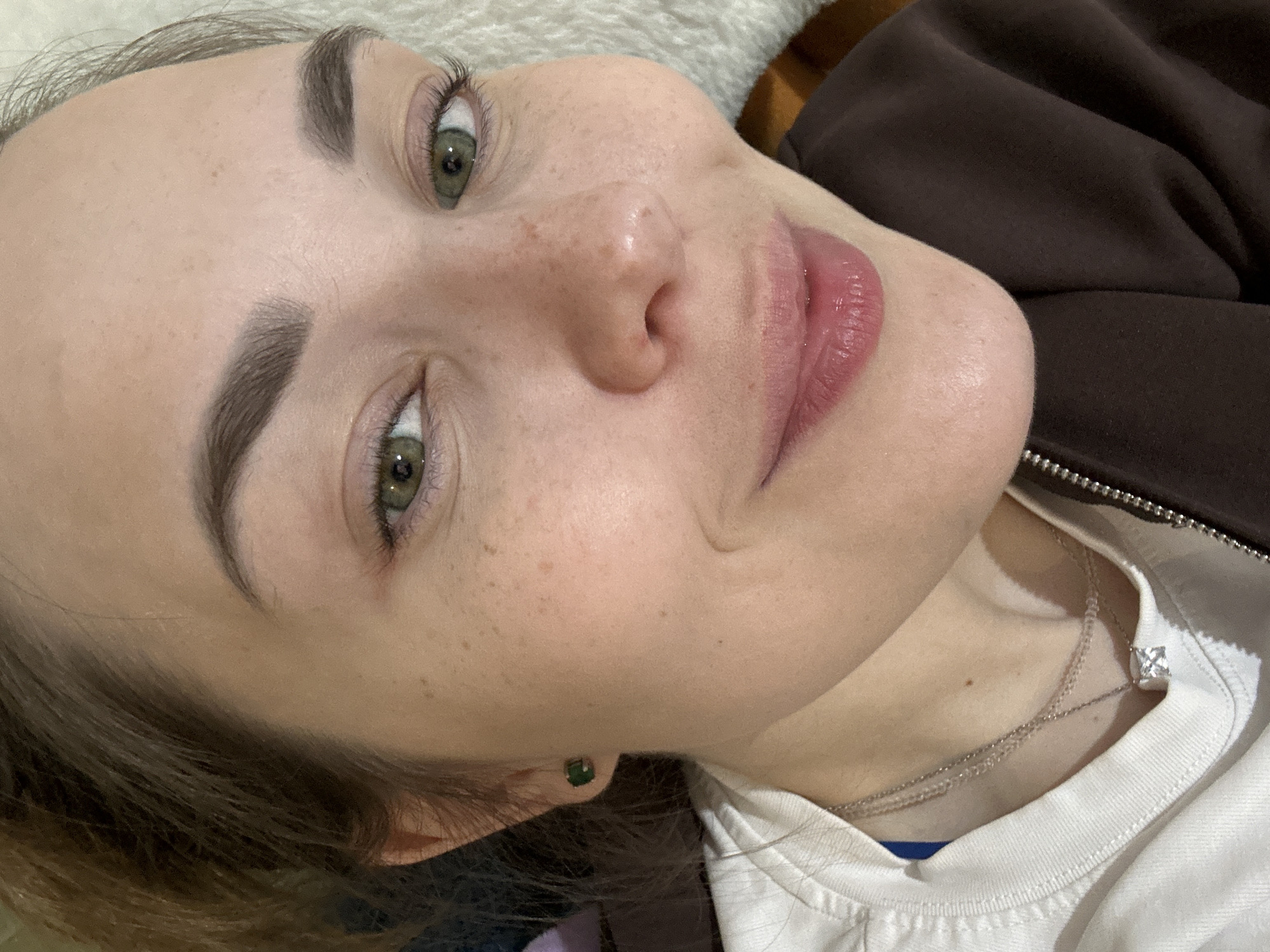 A close-up portrait shows a young woman from the mid-chest up, positioned at a slight tilt. She is lying down and looking directly at the camera with a calm, serene expression that suggests a subtle, gentle smile. Her eyes are a striking green, complemented by dark, well-defined eyebrows and visible eyelashes. Her fair skin features several small freckles across her nose and cheeks. Her lips are a natural pink, slightly parted.
She has dark brown hair, with strands visible near her face and around her neck. She is dressed in a white collared shirt, layered beneath a dark brown, zippered jacket. A delicate silver chain necklace with a prominent square-cut clear pendant rests on her collarbone, over the white shirt. A small, square green stud earring is visible in her left ear.
The background consists of a soft, light-colored material, likely a blanket or pillow, indicating an indoor setting. The lighting is soft and even, highlighting the subject's features. There is no visible text in the image. The provided context states the location is Perm, Russia.