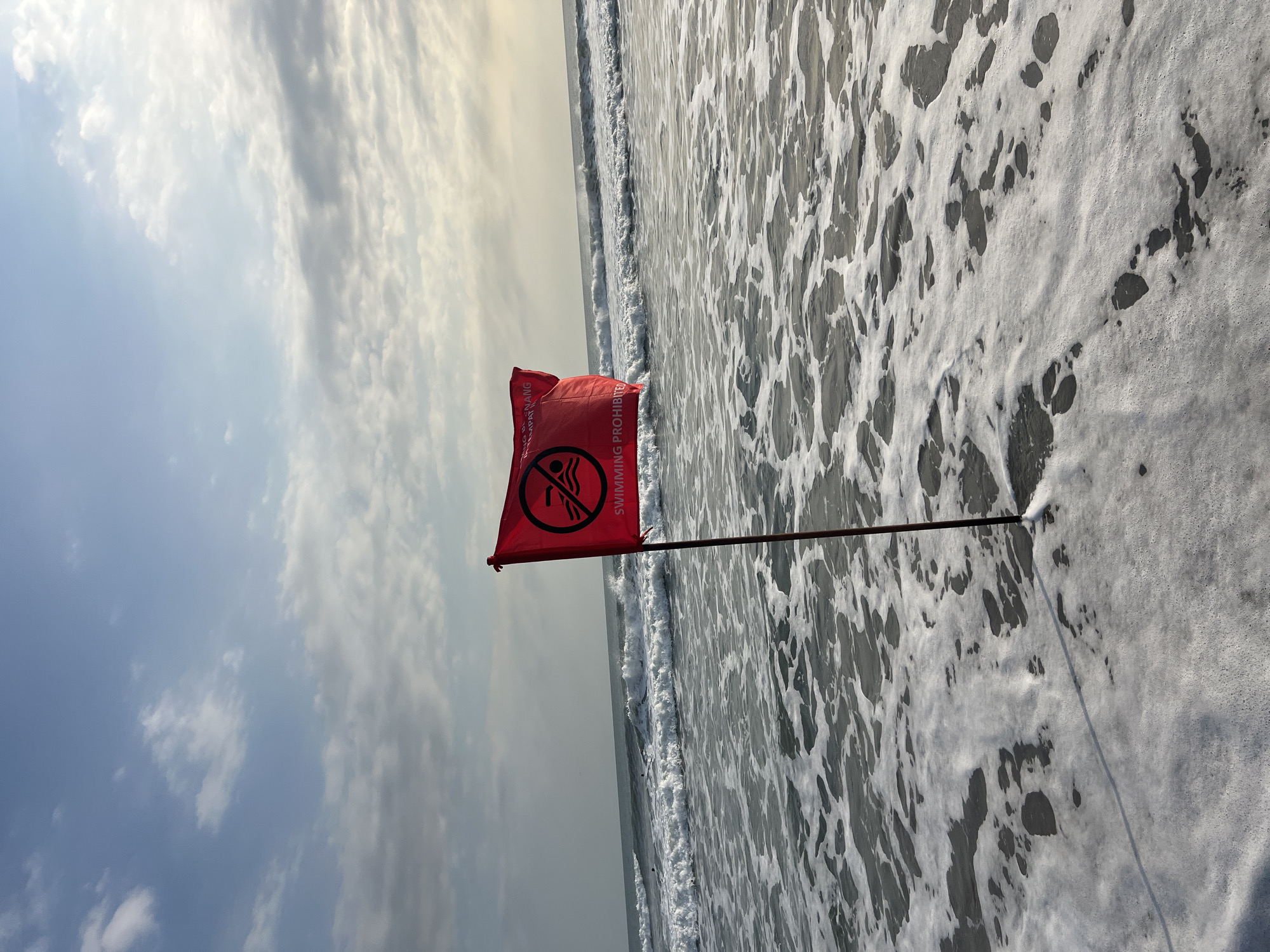 A red flag with a black circle and a symbol of a person swimming inside, crossed out, is planted in the shallow surf on a beach. Text on the flag reads "SWIMMING PROHIBITED." The flag is on a wooden pole and is partially submerged in the foamy water. Waves are breaking in the background, and the sky is partly cloudy. The scene appears to be the oceanfront in Kuta, Indonesia.