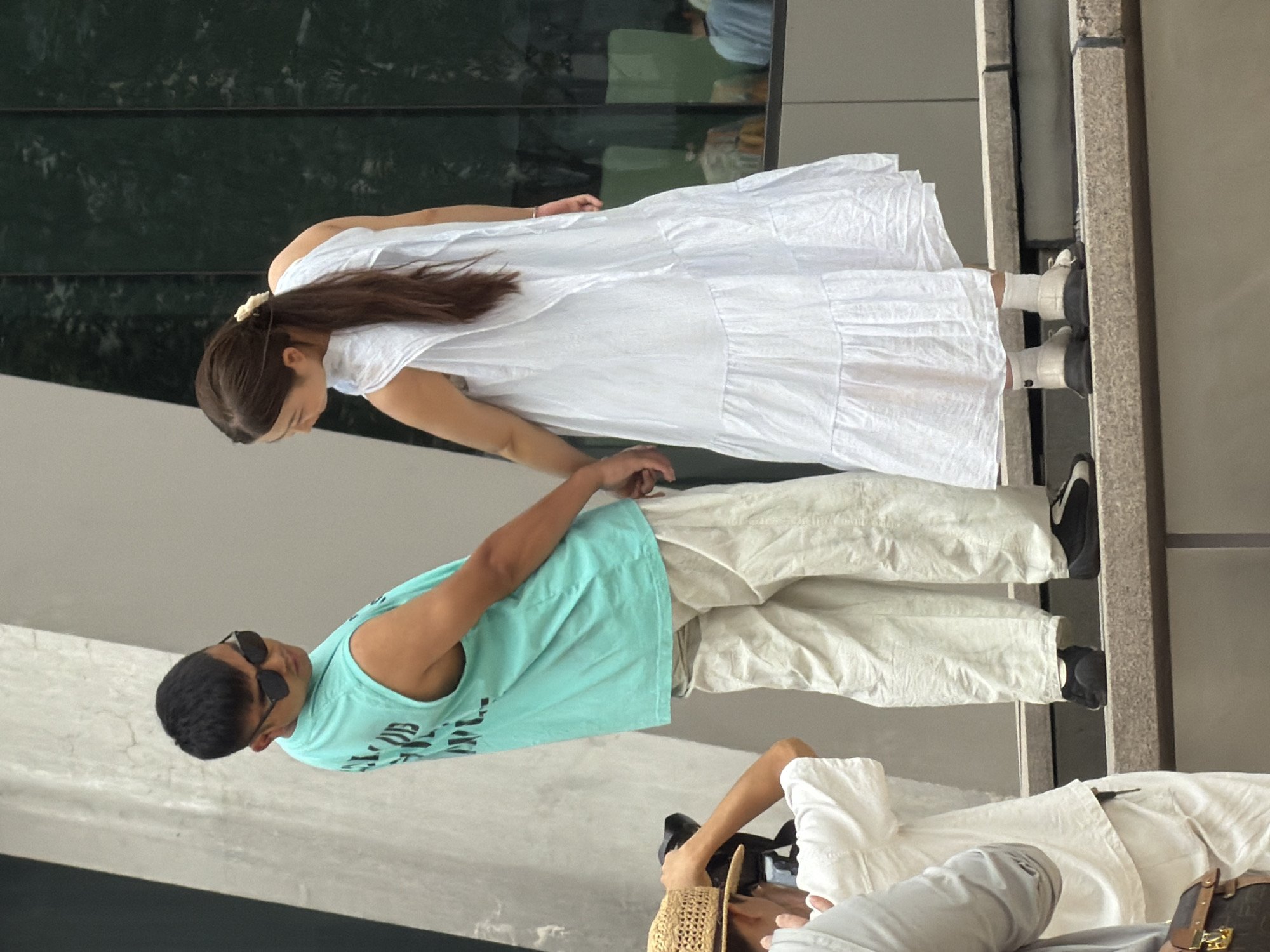 In Yuzhong District, China, a man wearing a turquoise tank top and light-colored pants, and sunglasses, stands with his hand linked with a woman wearing a white tiered dress. The man is facing away from the camera, looking to his left, while the woman is facing away from the camera, looking to her right. They are standing on a stone ledge in front of a building with a dark green reflective facade. To the left of the couple, a person wearing a straw hat and white clothing is visible holding a camera. Another person, wearing a light-colored shirt, is partially visible in the lower left corner. The background shows a large, modern building with some greenery reflected in its windows.