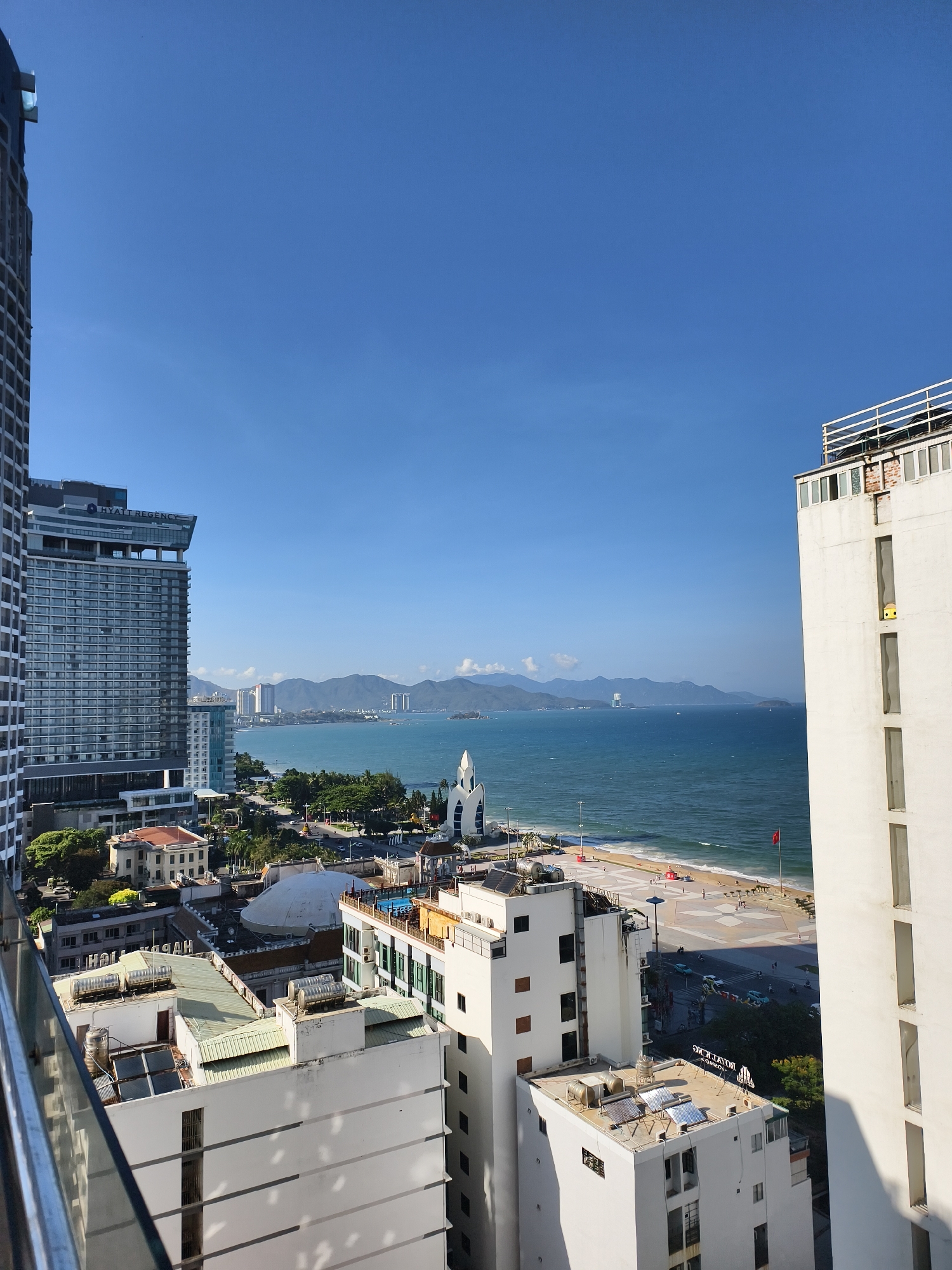 The image presents a cityscape, likely a coastal city in Southeast Asia. The sky is clear and blue. The view is from a high vantage point, possibly a hotel balcony. Tall buildings dominate the left and right foreground. A large hotel, labeled "Hyatt Regency", is partially visible on the left.
In the midground is the coastline, with a beach visible. A promenade runs along the beach, where a few pedestrians are present. A white, spire-like structure stands near the beach. Mountains are visible in the background across the sea.
The foreground shows parts of several buildings. The overall environment suggests a warm, sunny climate.