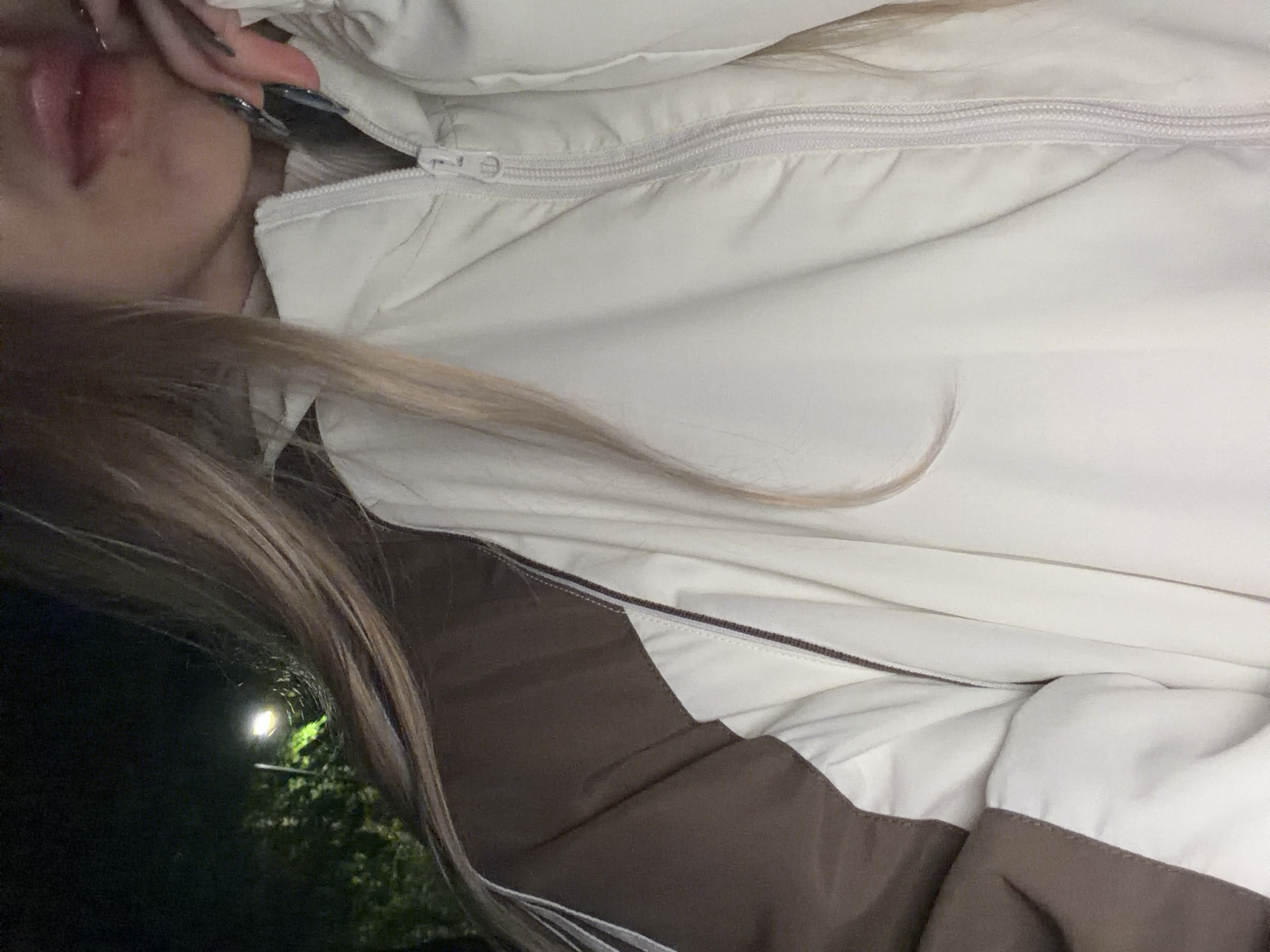 This image shows a person wearing a white jacket with a zipper closure. The jacket has brown accents along the lower section. A strand of light brown hair is visible across the jacket. The person's lips are slightly visible in the upper left corner. In the background, out of focus, there is a light post illuminating some trees. The location appears to be outdoors at night. The city is Unknown, Unknown.
