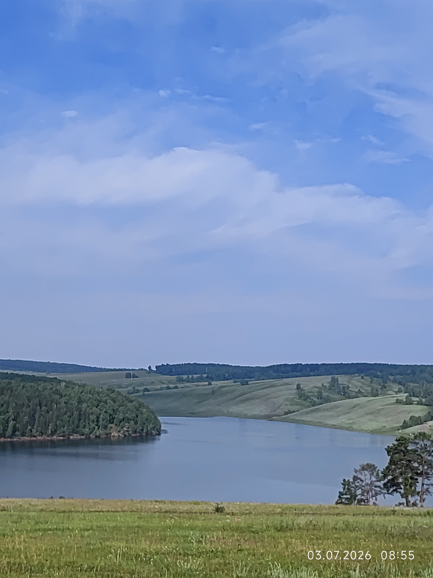 This is an aerial, landscape photograph of a river bend surrounded by rolling, grassy hills and a dense forest. The sky is blue with scattered white clouds. The foreground is a grassy field. In the lower right corner, the date "03.07.2026" and time "08:55" are superimposed. The location is Konovalovo, Russia.