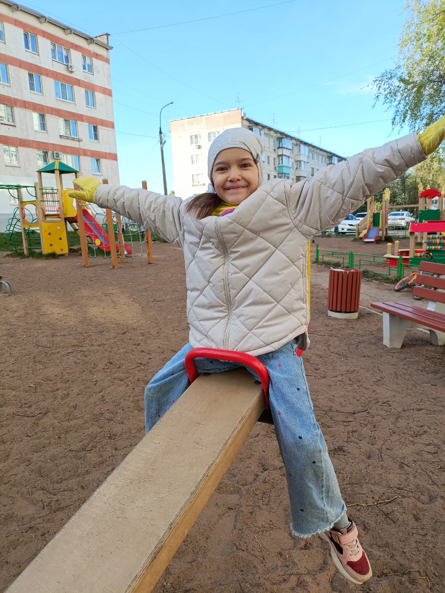The photograph shows a young girl, likely a child, sitting on a seesaw in a playground. She is wearing a light-colored quilted jacket, blue jeans adorned with heart designs, and pink sneakers. Her arms are outstretched, and she is smiling at the camera. On her head, she is wearing a grey beanie or headscarf and yellow gloves.
The background reveals a typical urban playground setting. There are several other playground structures visible, including slides and climbing frames. Behind the playground, multi-story apartment buildings can be seen, indicating a residential area. A bench and waste bin are also present near the seesaw. The ground of the playground is covered in sand.
Based on available information and visual cues, this image was taken in the city of Chaikovsky, Perm Krai, Russia.