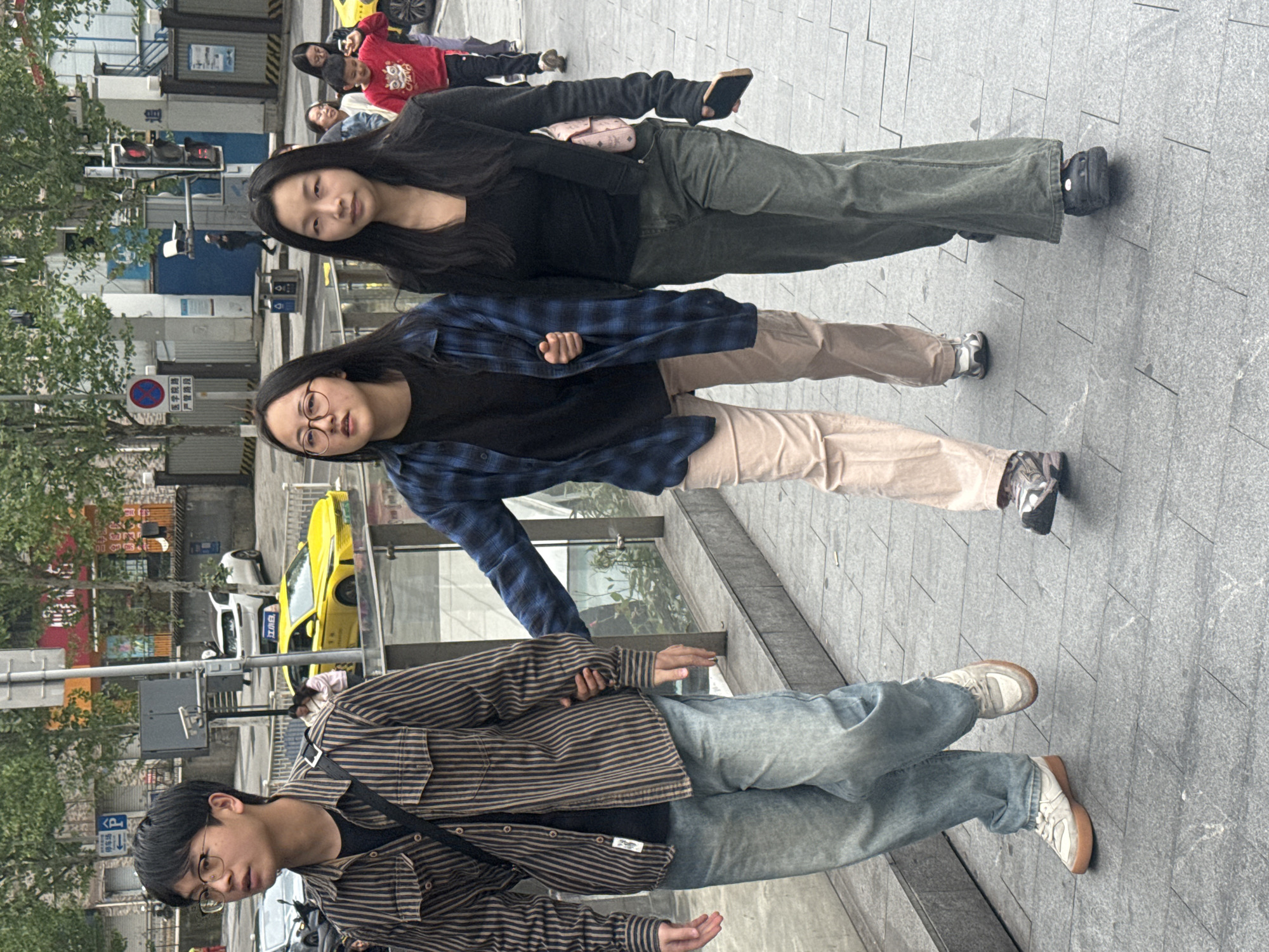Three individuals stand on a paved walkway. The first individual, a young man, wears glasses, a striped brown shirt, a black t-shirt underneath, blue jeans, and white sneakers. He has a shoulder bag. The second individual, a young woman, wears glasses, a black t-shirt, a blue and black plaid shirt, and light tan pants. The third individual, a young woman, wears a black shirt and olive green pants. In the background, there is a yellow taxi, buildings, trees, and street signs. The location appears to be an urban area in the Yuzhong District of China.