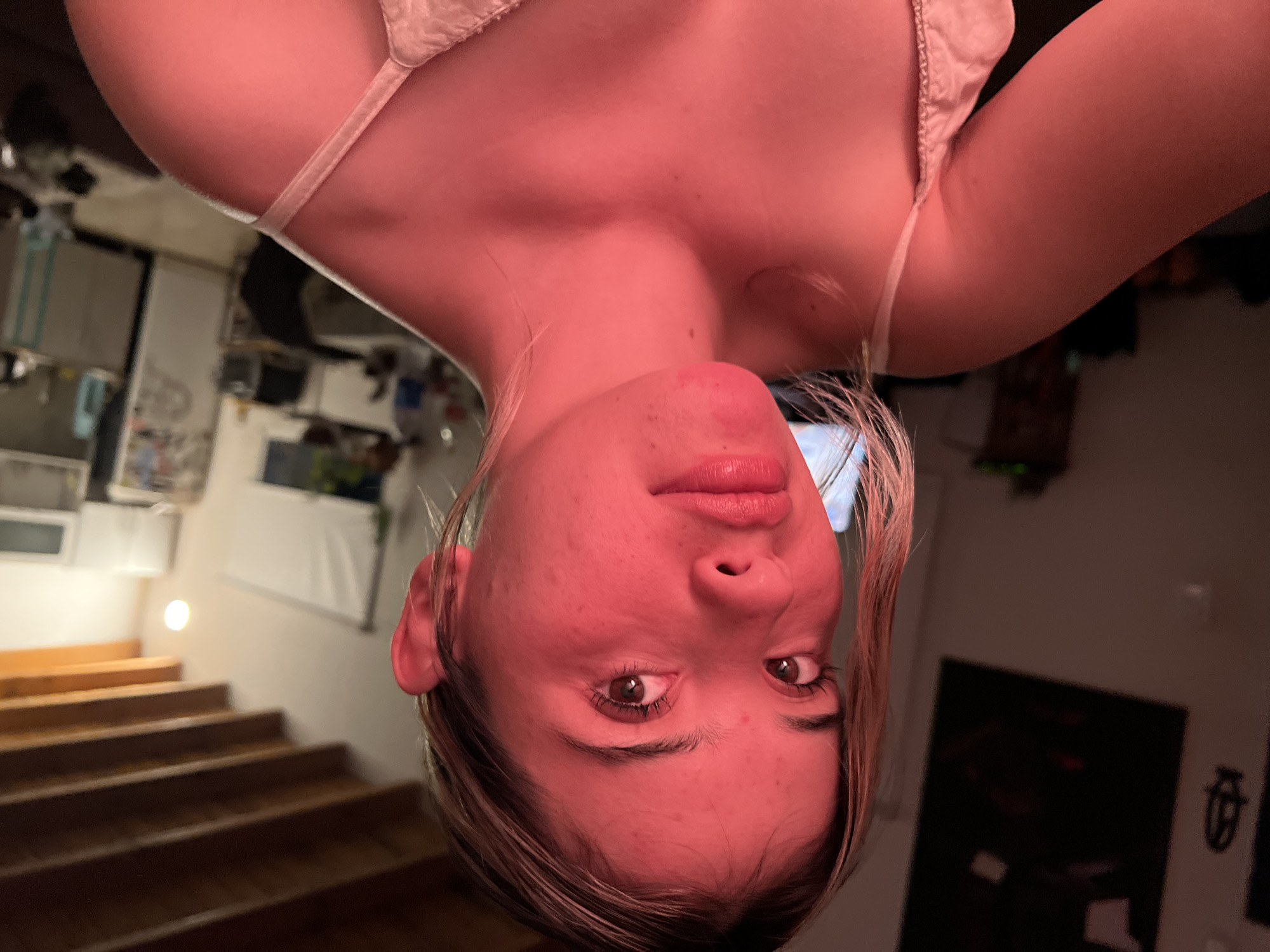 A medium close-up shot shows a young woman with light brown hair illuminated by a strong red light source. She is looking directly at the camera with a neutral expression. Her face is slightly flushed, and there are some subtle blemishes visible. She is wearing a light-colored camisole with thin straps. The background is out of focus and appears to be an indoor setting, possibly a living room or kitchen, with wooden ceiling beams visible at the top of the frame. The overall lighting suggests it is nighttime. The exact location in Sumalao, Argentina, cannot be determined from the visual information alone.