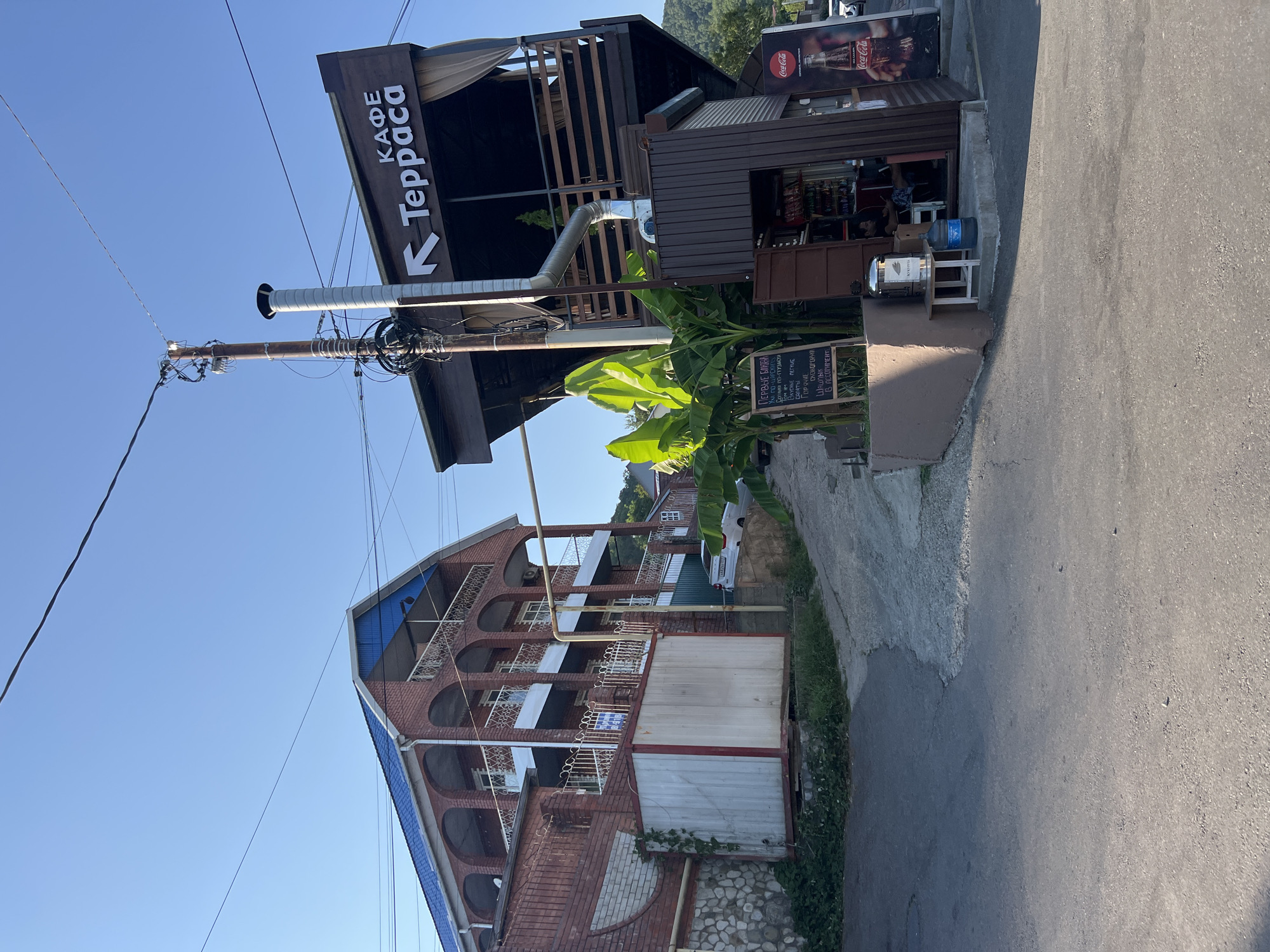 This image depicts a street scene in Tuapse, Russia, on a clear, sunny day. On the right side of the frame, a two-story establishment with a dark brown, angled wooden facade is visible, featuring outdoor seating areas. A prominent sign with a white arrow and the word "Терраса" (Terrace) in white Cyrillic letters is mounted on the upper story. Below the outdoor seating, a small kiosk-like structure is open, with a person seated inside. To the left of this establishment, a large container, possibly a storage unit, is positioned against a brick building with arched balconies. A paved road occupies the foreground, with a gravelly incline leading up towards the buildings. Lush green banana plants are growing near the cafe. A Coca-Cola advertisement poster is affixed to the side of the cafe. The overall impression is a commercial area with residential elements, set against a backdrop of hills or mountains on the right side of the frame.