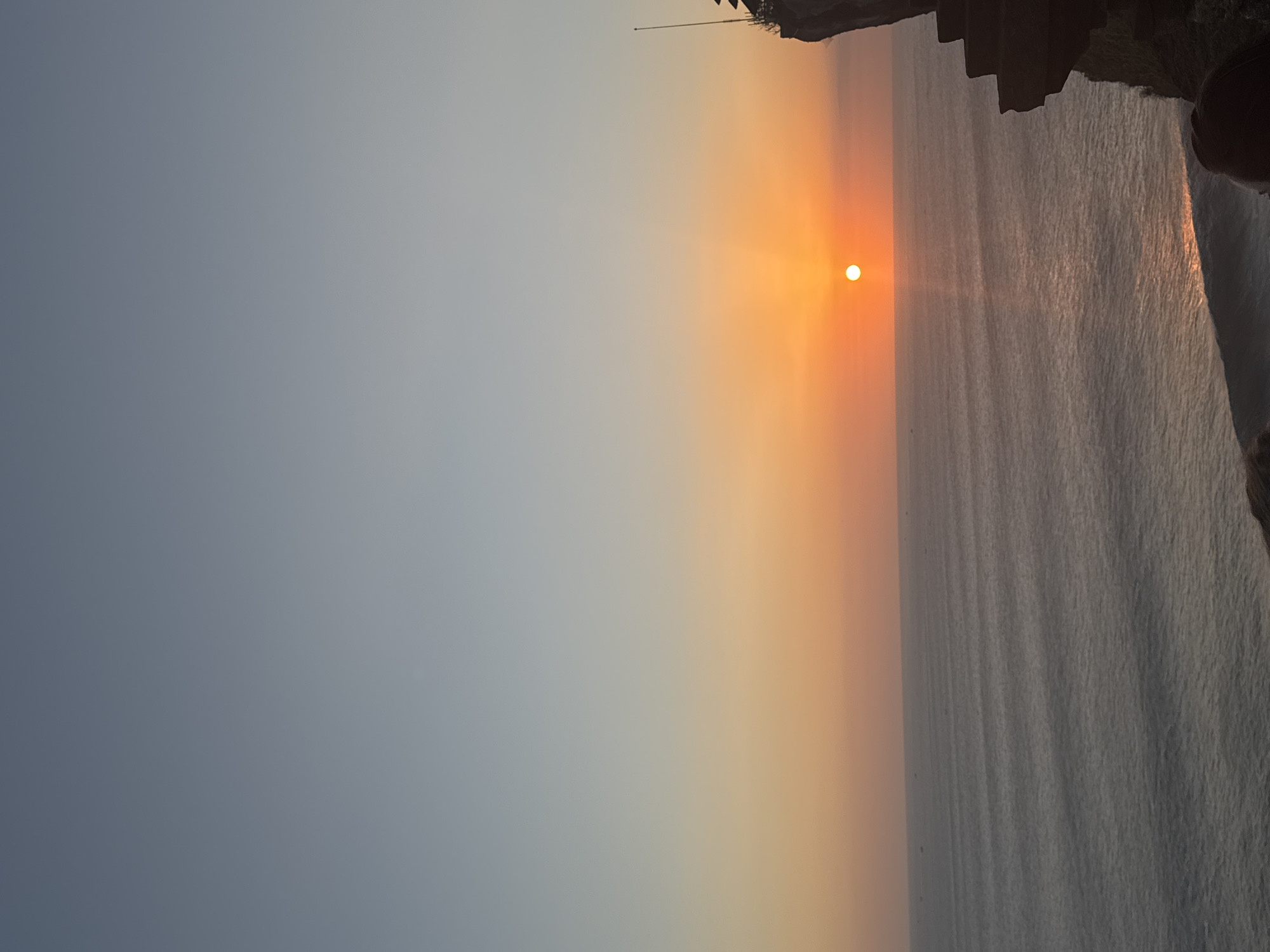 The image shows a sunset over the ocean. The sun is a bright orange orb low in the sky, casting a golden reflection on the water. The ocean surface has gentle waves, and there are small boats visible on the horizon. The sky transitions from a pale blue at the top to a warm orange near the horizon. On the right side of the image, a dark, rocky cliff face is visible, with some man-made structures on top. The scene appears to be a scenic overlook.