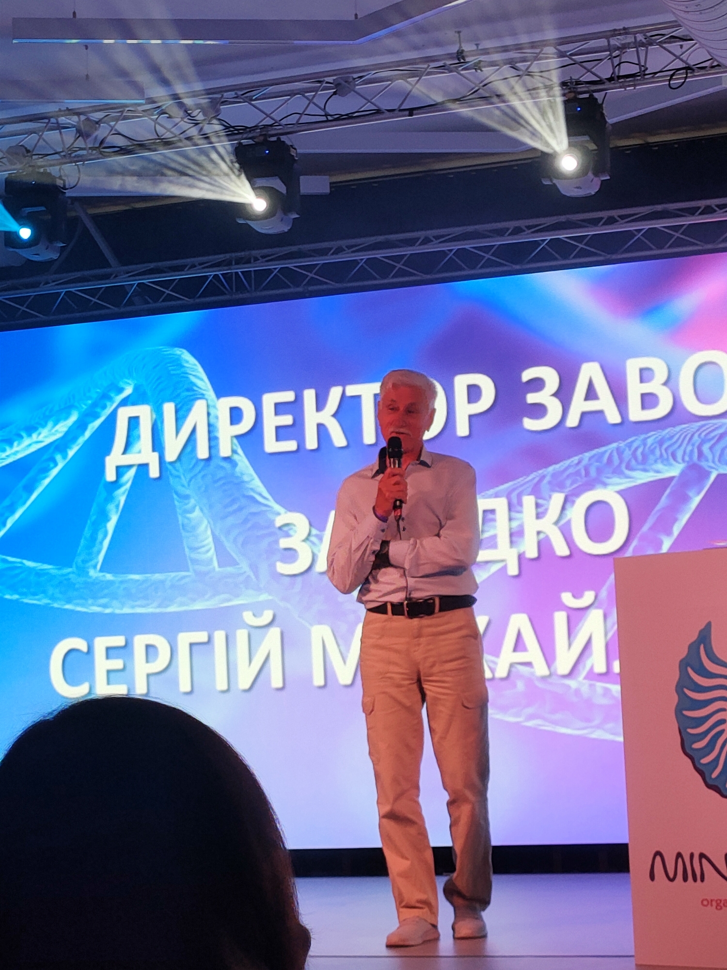 A man with gray hair and a light blue button-down shirt and tan cargo pants is speaking into a microphone on a stage. He is wearing a black belt. Behind him is a large screen displaying a blue and purple background with white text that appears to be in Cyrillic. There are also DNA strand graphics on the screen. To the right of the man is a white panel with a blue logo and text that reads "MIN" in large font and smaller text below it. The stage is lit by spotlights from above. The overall scene suggests an event or presentation taking place in a venue. Based on the text on the screen and the logo, it is likely an event related to a company or organization. This scene is likely from Kyiv, Ukraine.