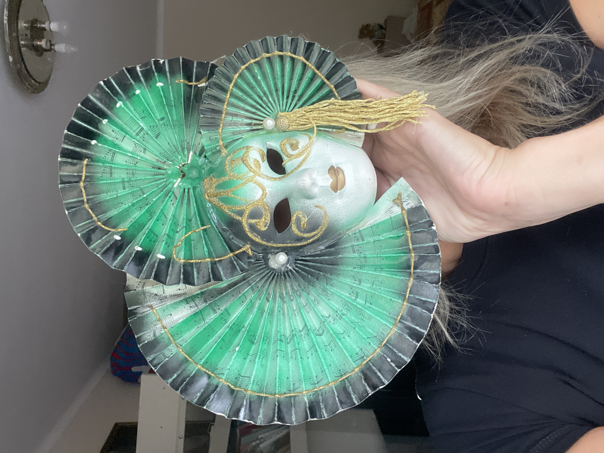 A person holds a decorative mask. The mask has a white face with gold detailing around the eyes and nose, and it's surrounded by green and black fan-like structures with gold trim. Music notes are printed on the green parts of the fan. There is a gold tassel on top of the mask. The background shows a room interior with a wall sconce and a person wearing a black shirt. The location is not identifiable as Sochi, Russia from the image.