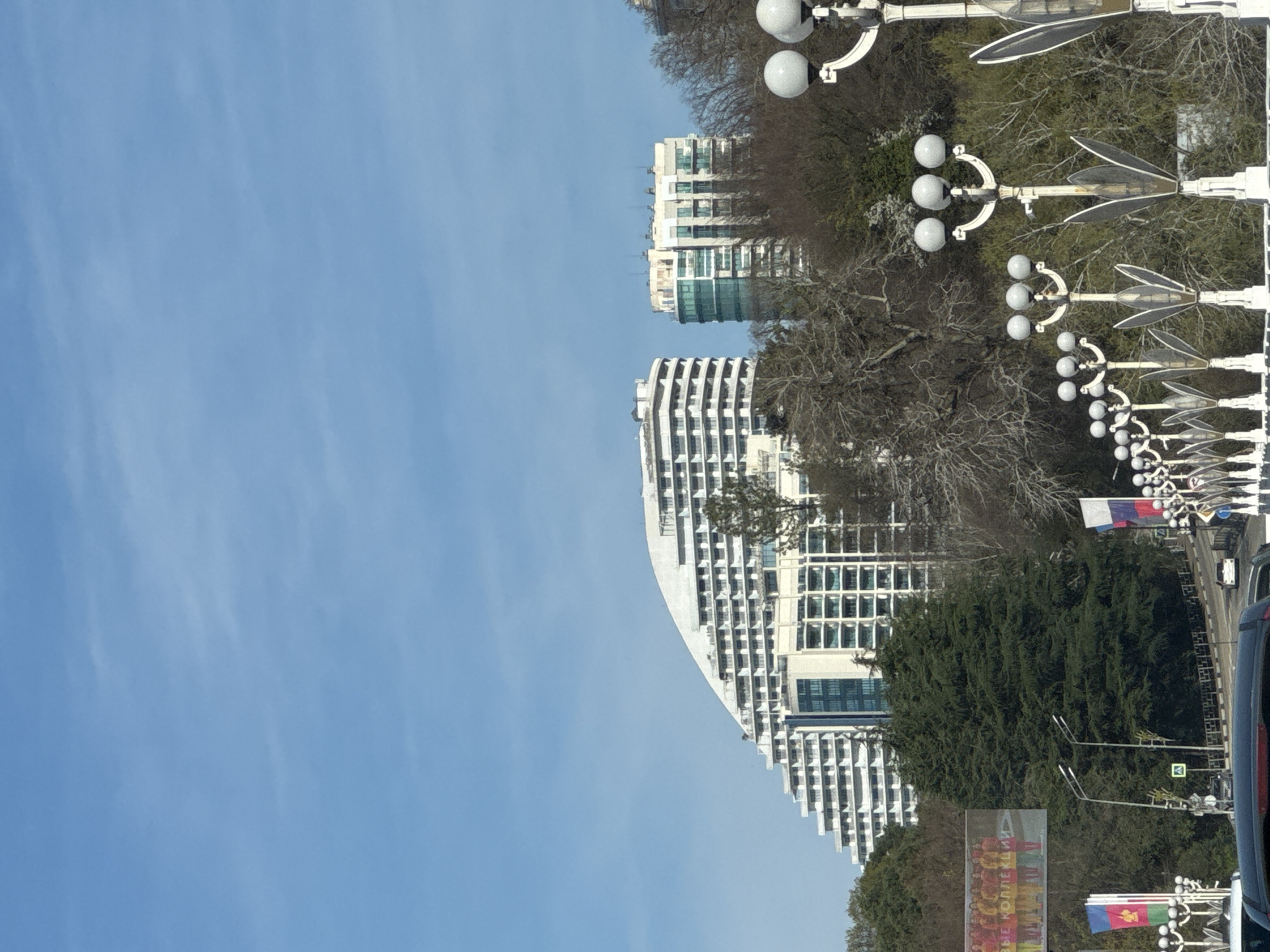 The image captures a street view in Sochi, Russia. Prominent features include modern, multi-story white buildings with a curved design. A row of decorative street lamps lines the road, and trees are interspersed between the buildings and the lamps. The sky is clear blue. In the foreground, a road with vehicular traffic is visible, along with flags.
