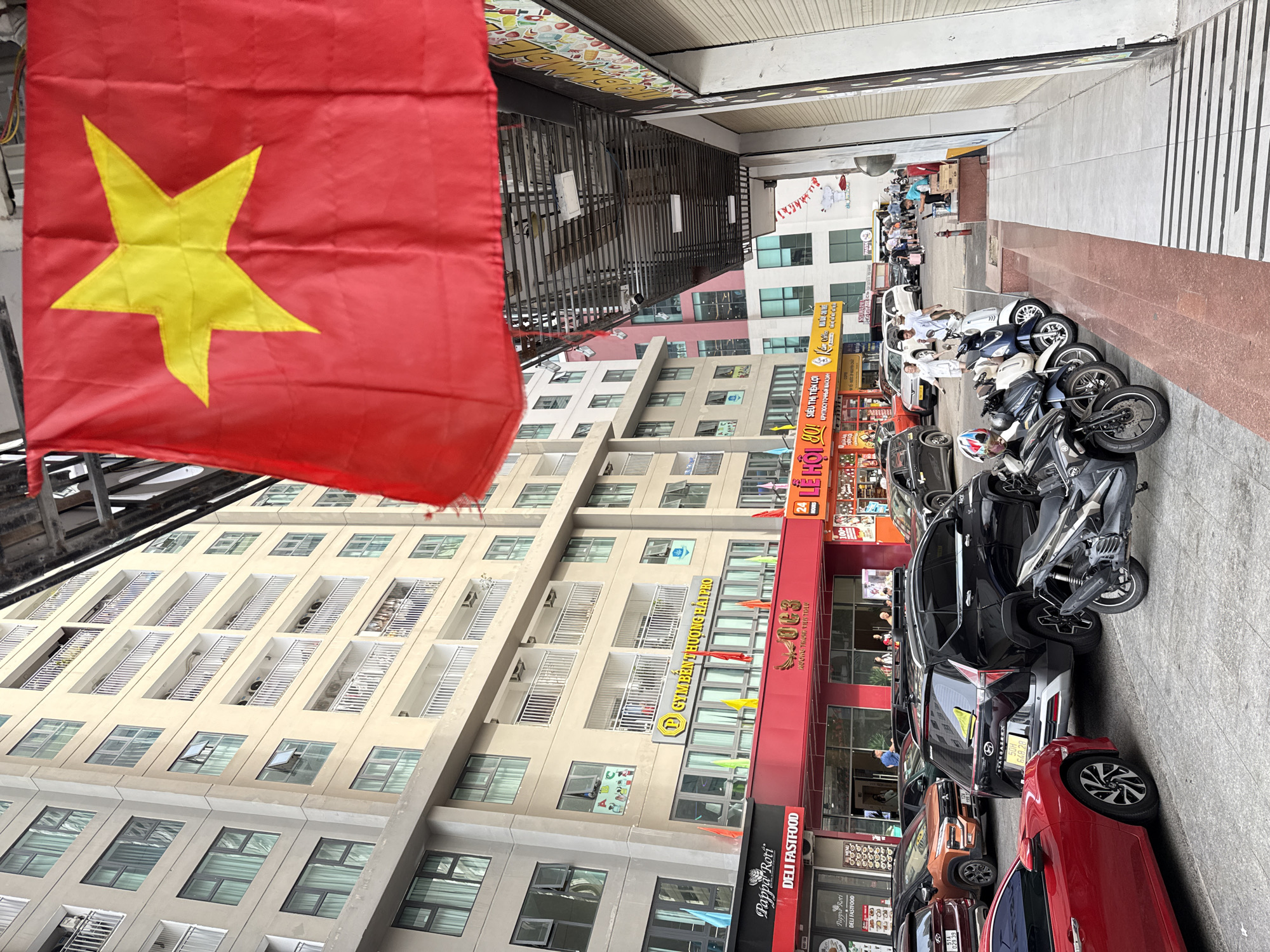 The image depicts a street scene in Nha Trang, Vietnam, with a focus on a modern, multi-story building and the street in front of it. A large Vietnamese flag, predominantly red with a yellow star, is prominently displayed in the upper right portion of the frame, partially obscuring the building. The building itself appears to be a residential or commercial complex, with numerous windows and balconies.
The ground level of the building features several businesses with awnings and signage. One sign reads "Pappa Roti DELI FASTFOOD." Another business has signage that appears to include Vietnamese text and a logo with a star. Various flags and banners are hung along the storefronts.
The street in front of the building is lined with parked vehicles, including cars and numerous motorbikes, suggesting a busy urban environment. A red car is parked in the foreground to the left. Several people are visible in the background, some walking and others near the storefronts. The overall impression is of a commercial street in a Vietnamese city, possibly during daylight hours given the lighting. The architecture and vehicles are consistent with a modern urban setting in Vietnam.