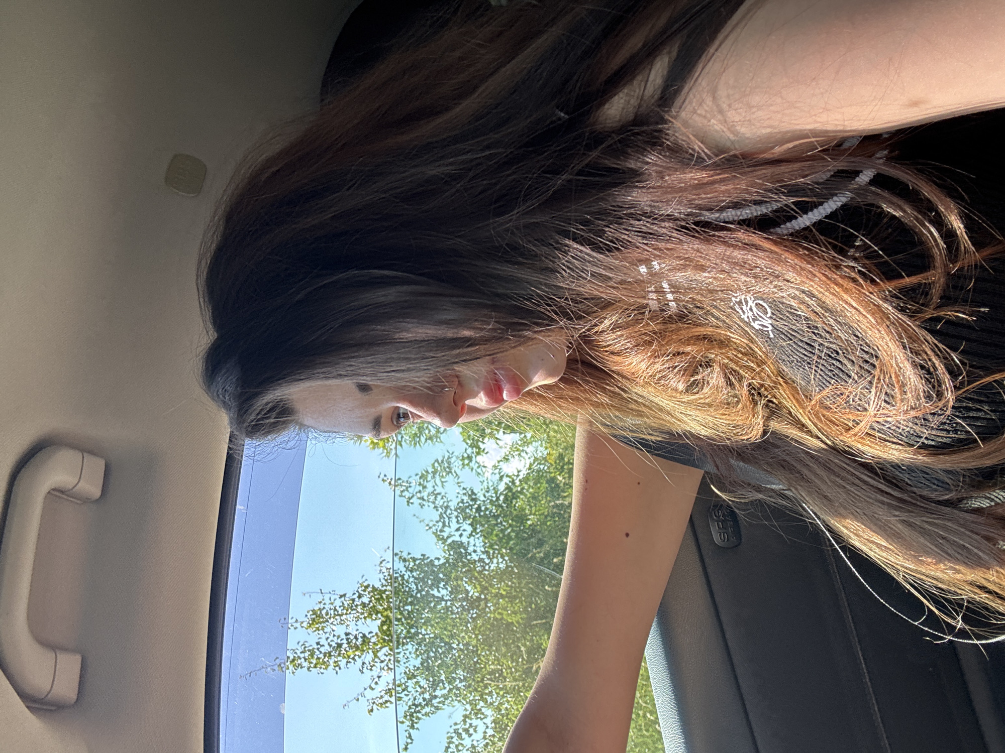 A young woman with long, dark brown hair is looking out the window of a car. She is wearing a black top with white trim. The car interior is visible, including a grab handle on the ceiling and the door panel with an "SRS AIRBAG" label. Outside the window, green trees and a blue sky are visible. The image appears to have been taken during the daytime, with sunlight illuminating the woman's hair. The location is not specified in the image or provided information.