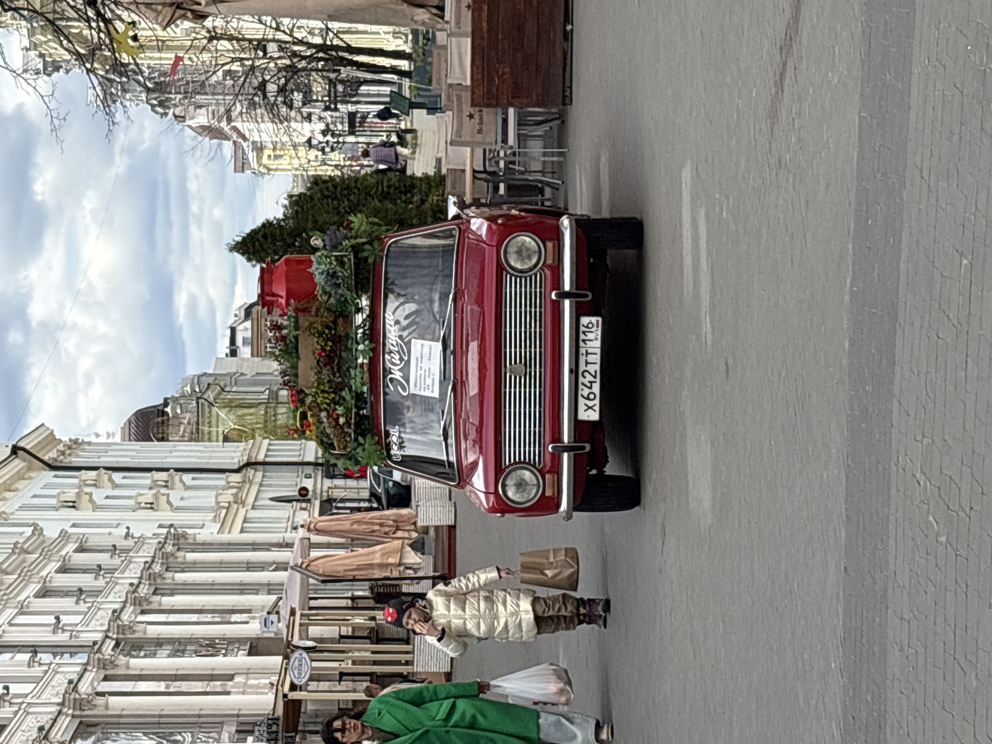 The image shows a street scene in Kazan, Russia. In the foreground is a red vintage car adorned with plants on its roof. A person in a green coat and a person in a white puffer jacket walk along the sidewalk near the car. The sidewalk is paved with grey bricks. Tall buildings with detailed facades line the street. In the background, trees are visible. There is outdoor seating for a café. The car's license plate reads "X642TT 116".