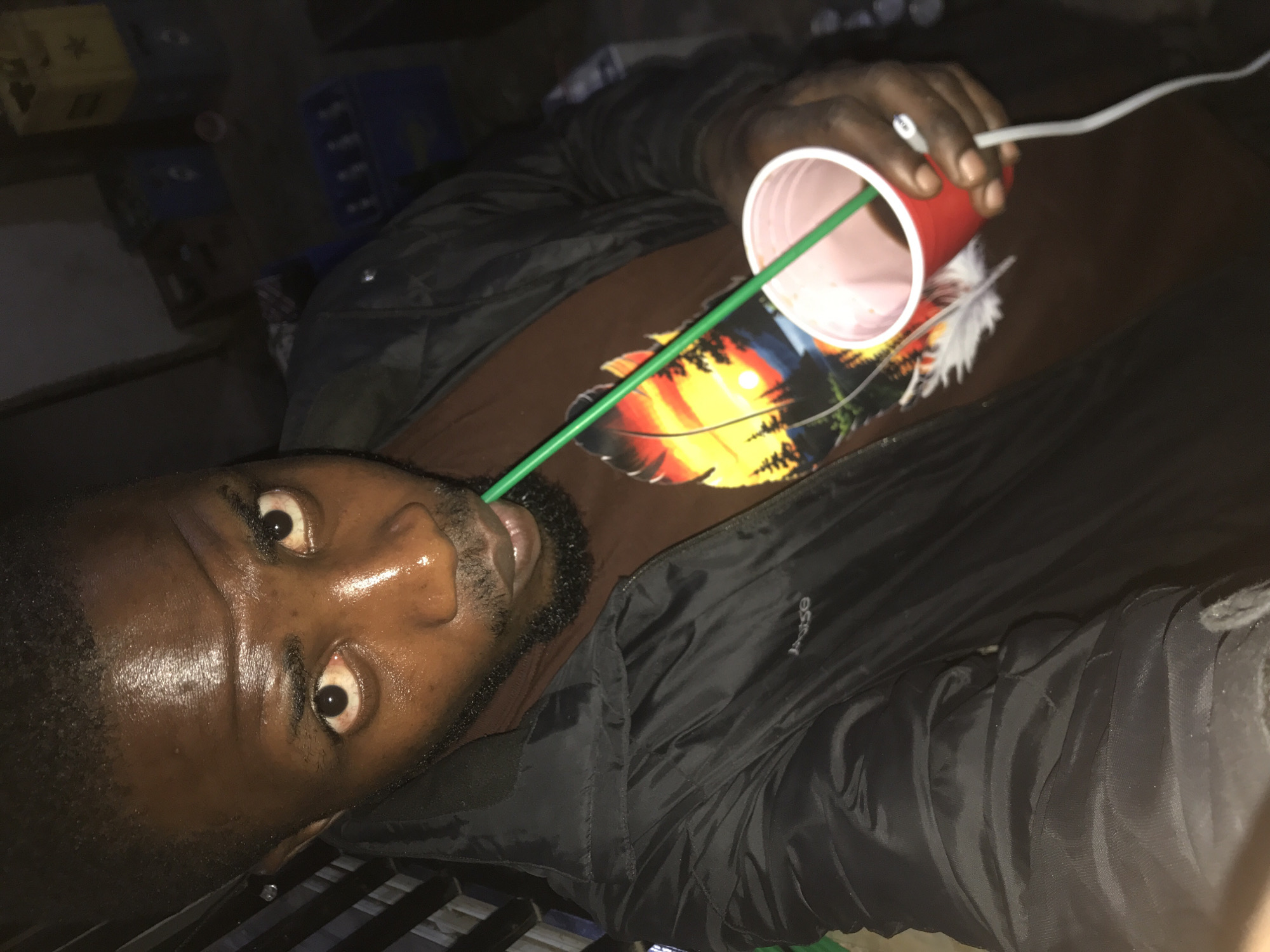 A dark-skinned man with a beard is shown in a close-up, selfie-style shot. He is wearing a black jacket over a brown t-shirt with a graphic design. He is holding a red plastic cup in his right hand, and a green straw is visible in his mouth, extending into the cup. His eyes are open, and his face appears slightly sweaty. The background is dark and appears to be an indoor or semi-outdoor space with some boxes and possibly shelving visible. The man is the primary subject of the image.