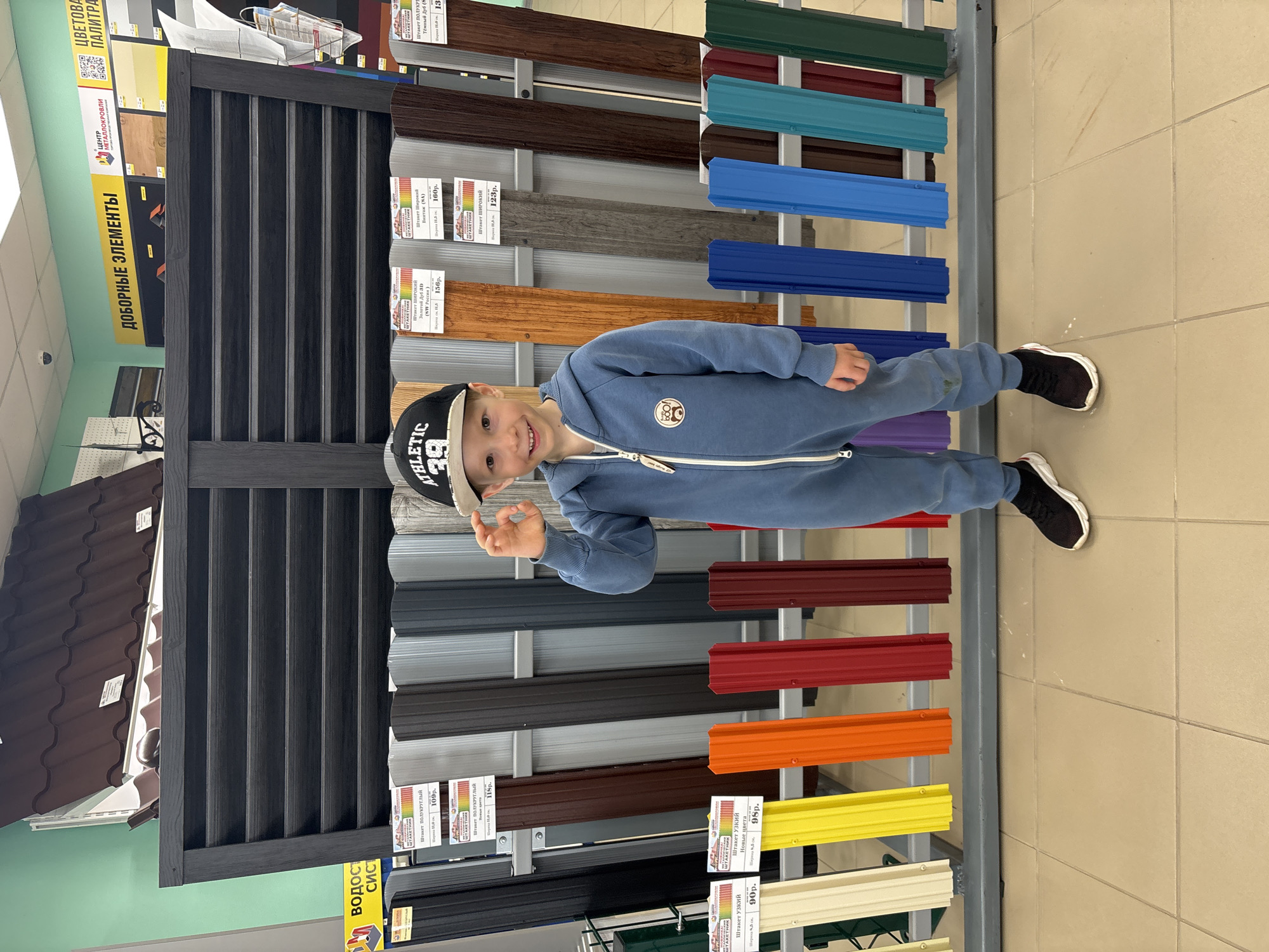 A boy in a blue jumpsuit stands sideways in front of a display of colored metal roofing materials in what appears to be a hardware store. The boy is wearing a black and white baseball cap with the word "ATHLETIC" and the number "39" printed on it. He is smiling and making an "okay" gesture with his hand. The roofing materials are arranged vertically in various colors, including brown, gray, blue, red, orange, and yellow. Price tags are visible on each material. The background includes a tiled floor and some additional store displays. Text on the display materials reads "ДОБОРНЫЕ ЭЛЕМЕНТЫ" (Additional Elements) and "ЦВЕТОВА ПАЛИТРА" (Color Palette).
The scene may be in Chaikovsky, Russia, according to text visible on the displayed materials.