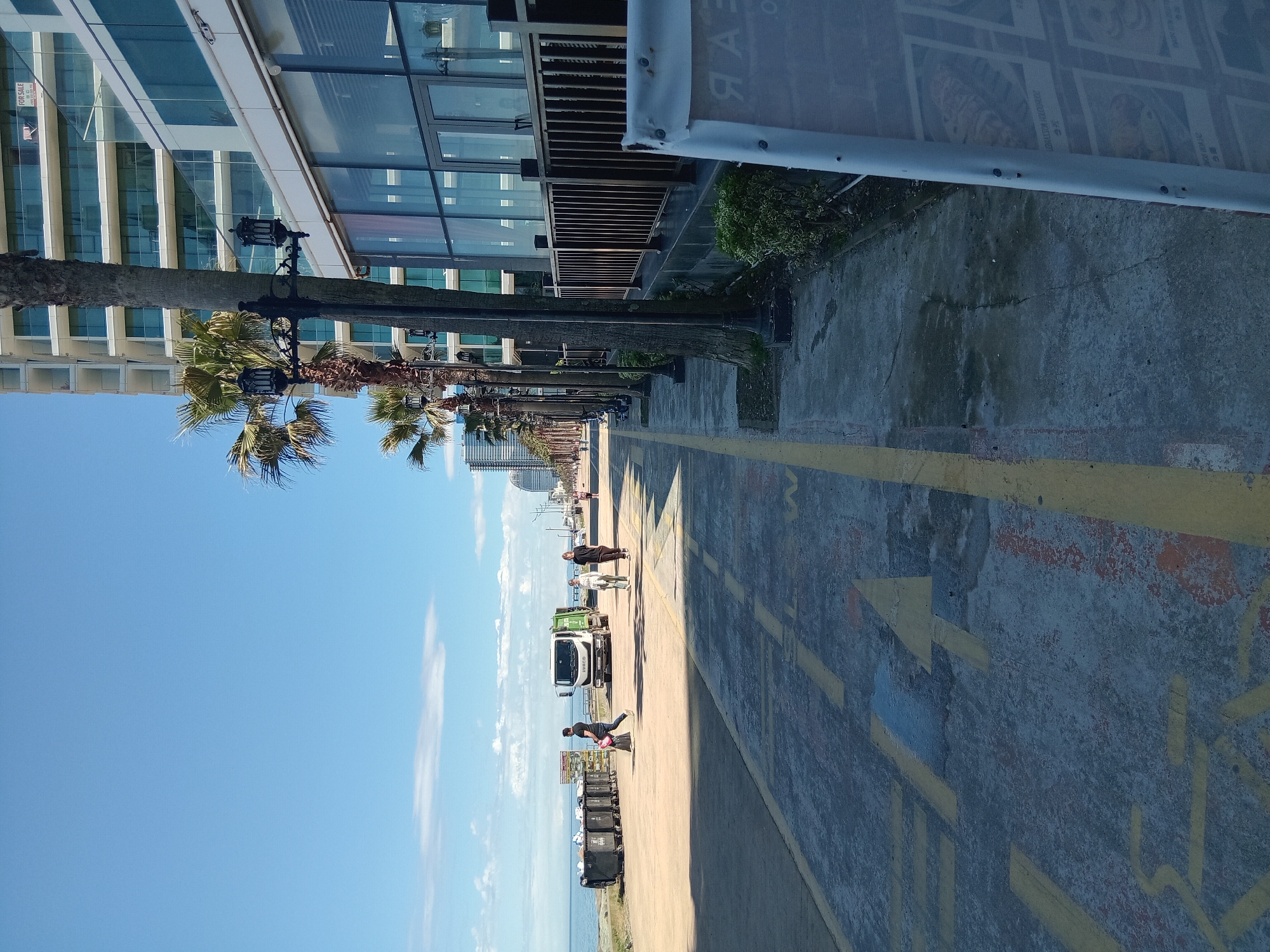 The image shows a street scene in Batumi, Georgia. It is a sunny day with a clear blue sky and a few scattered clouds. The street is lined with palm trees, streetlights, and modern buildings with glass facades. A sidewalk runs parallel to the street, marked with yellow lines and painted with yellow directional arrows. Several people are walking along the street, and a white truck is parked nearby. There are also black trash cans along the seaside. The Black Sea is visible in the background.