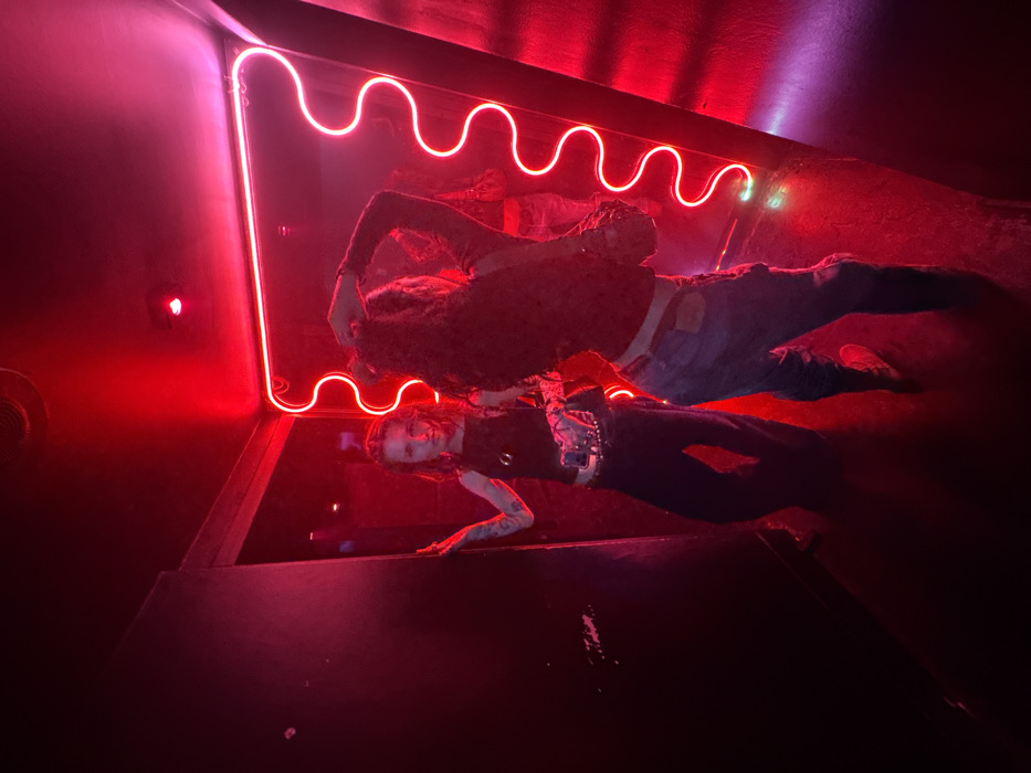 Two individuals are reflected in a mirror under red lighting. Above the mirror, a wavy red neon light fixture is visible. One person is wearing dark clothing and appears to be reaching out with their hand. The other figure is in jeans and a darker top, laying further back. The overall scene has a dark and somewhat distorted aesthetic due to the lighting and the mirror's reflection.