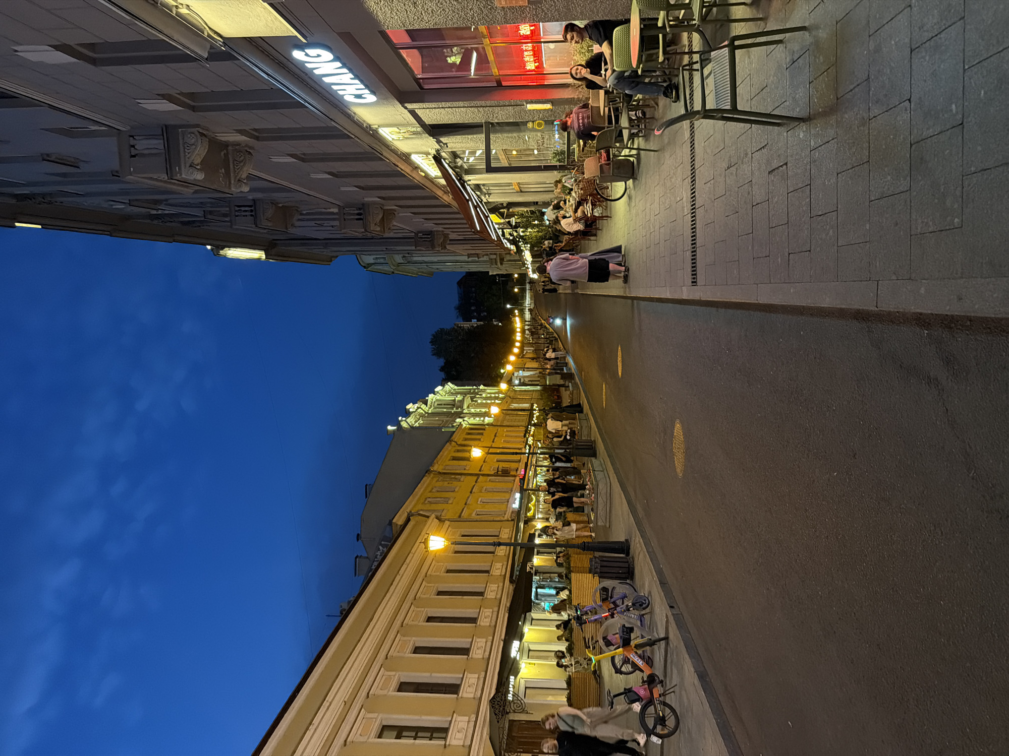 The image depicts a street scene in Moscow, Russia, during twilight hours. The street is lined with two-story buildings with illuminated windows and storefronts, suggesting commercial activity. On the left side of the street, a row of yellow buildings with traditional architectural elements such as decorative cornices and arched windows is visible. Several outdoor seating areas with umbrellas are set up in front of restaurants and cafes. On the right side, a modern building features a prominent sign that reads "CHANG" in illuminated lettering.
People are scattered along the sidewalks, some walking and others seated at outdoor tables, engaging in conversation and dining. Several bicycles and electric scooters are parked along the sidewalk. The street itself appears to be paved with asphalt, and streetlights cast a warm glow on the scene. The sky is a deep blue, indicating the transition from day to night, with a few wispy clouds present. The overall atmosphere suggests a lively urban evening with people enjoying outdoor amenities.