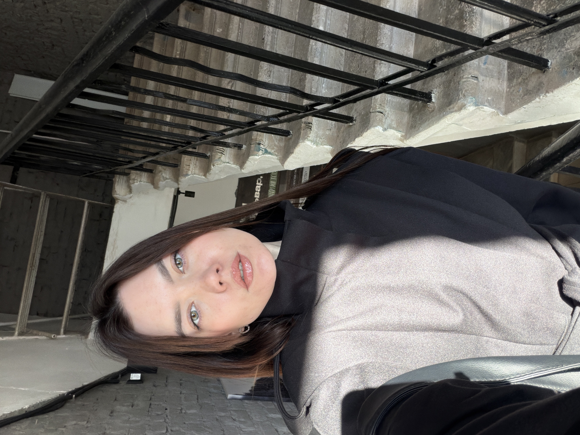 A young woman with brown hair and green eyes is posing in a photo with a metallic structure and a textured wall as a backdrop. She is wearing a gray jacket with a black collar, and what appears to be a black leather bag or accessory is visible at the bottom of the frame. The photo seems to be taken outdoors in an urban environment.
While the location is specified as Chaikovsky, Russia, without additional contextual information or specific landmarks, it's impossible to pinpoint the exact spot. More details would be needed to provide a precise identification of the location.