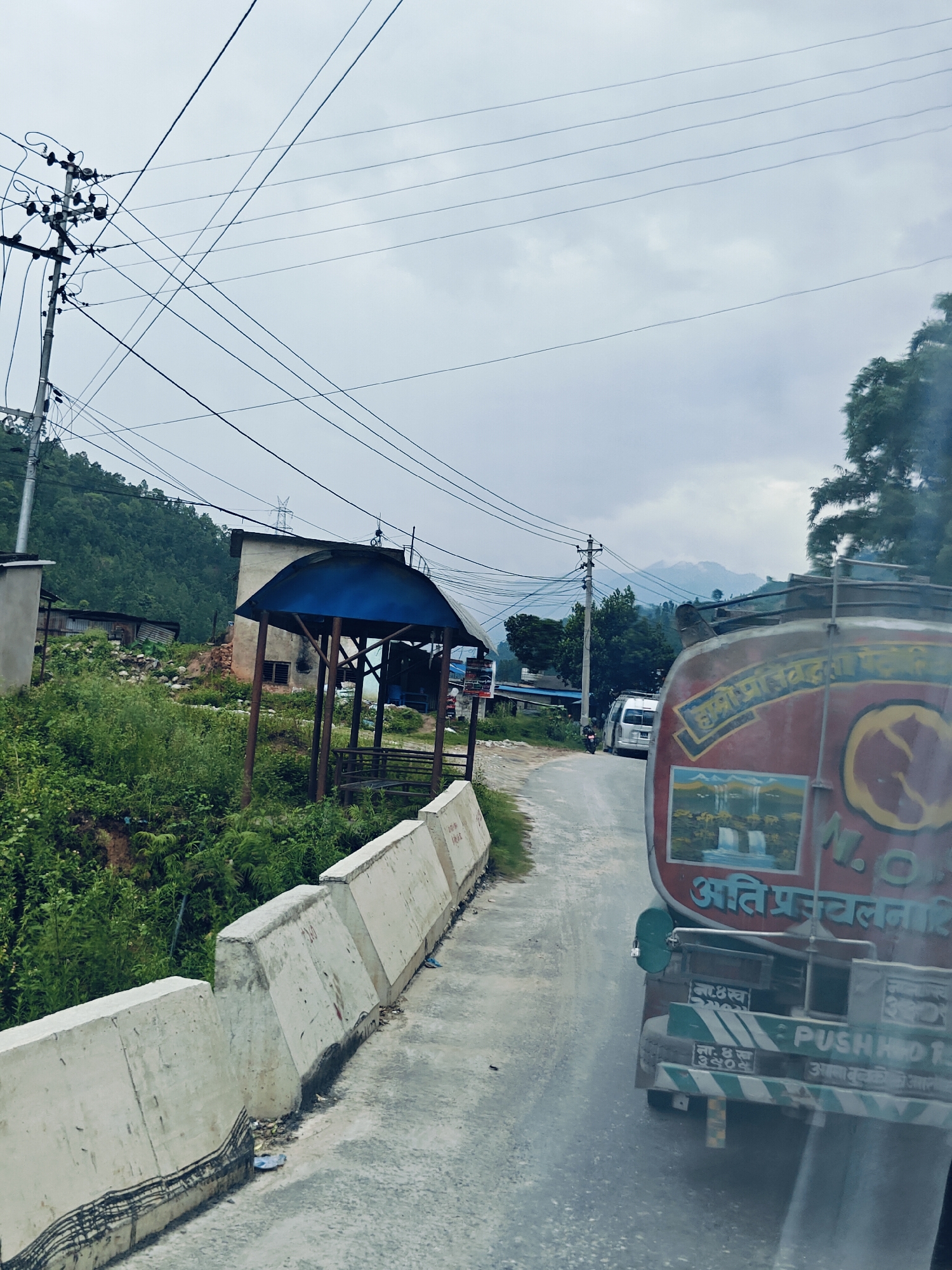 The image captures a street scene in Galchhi, Nepal, under an overcast sky. A truck, painted red and adorned with a depiction of waterfalls and text in Nepali, is prominently featured on the right side of the road. The truck appears to be moving forward. The road itself is paved and curves slightly to the left. To the left of the road, a concrete barrier lines the edge, with dense green vegetation and a small structure with a blue roof beyond it. In the background, a hillside covered in lush green trees rises towards the cloudy sky. Power lines are strung across the scene, supported by utility poles. Further in the distance, mountains can be seen peeking through the clouds. A white van is parked on the side of the road near the green vegetation.