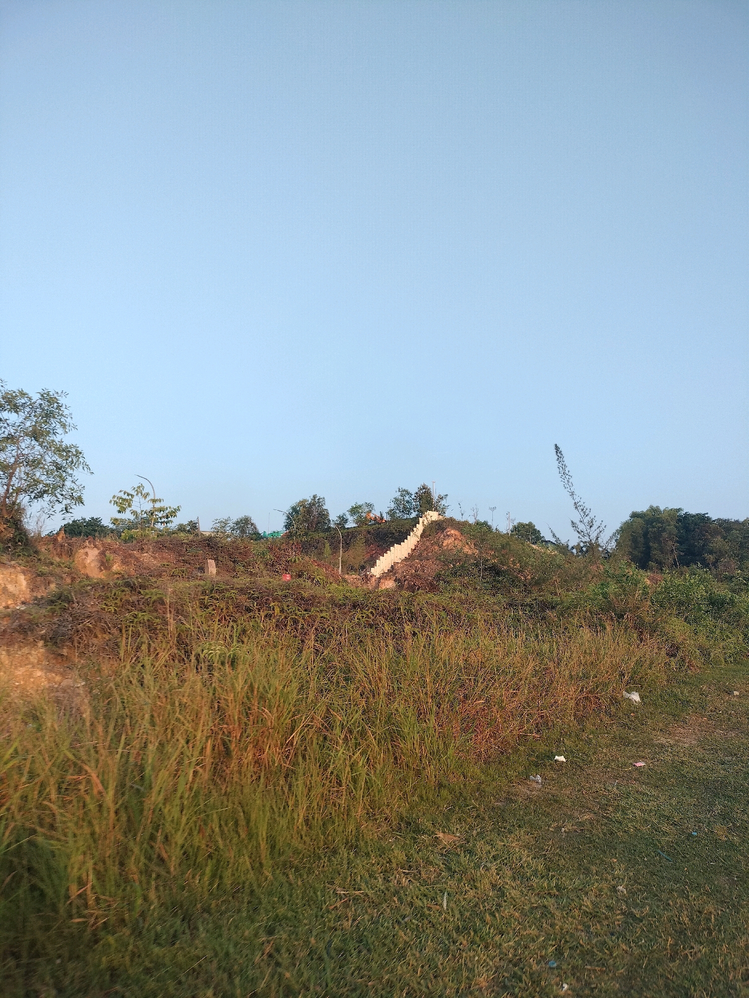 The image depicts an outdoor scene, likely a hillside or elevated area, with a clear, pale blue sky dominating the upper portion. The foreground is filled with tall, dry grass and some scattered green vegetation, suggesting a slightly unkempt or natural terrain.
In the middle ground, a man-made staircase with white concrete steps winds its way up a reddish-brown, earthen slope. This slope is covered with sparse vegetation and some exposed soil. Various trees and bushes are visible on the crest of the hill and in the background, forming a line of greenery.
To the far right of the image, a tall, dark, slender structure resembling a crane or antenna juts upwards into the sky. The overall impression is of an undeveloped or semi-developed area with some infrastructure, such as the staircase and the tall structure, present. The lighting suggests it might be late afternoon or early morning, given the soft light and long shadows that are not clearly visible. No people are visible in the image.