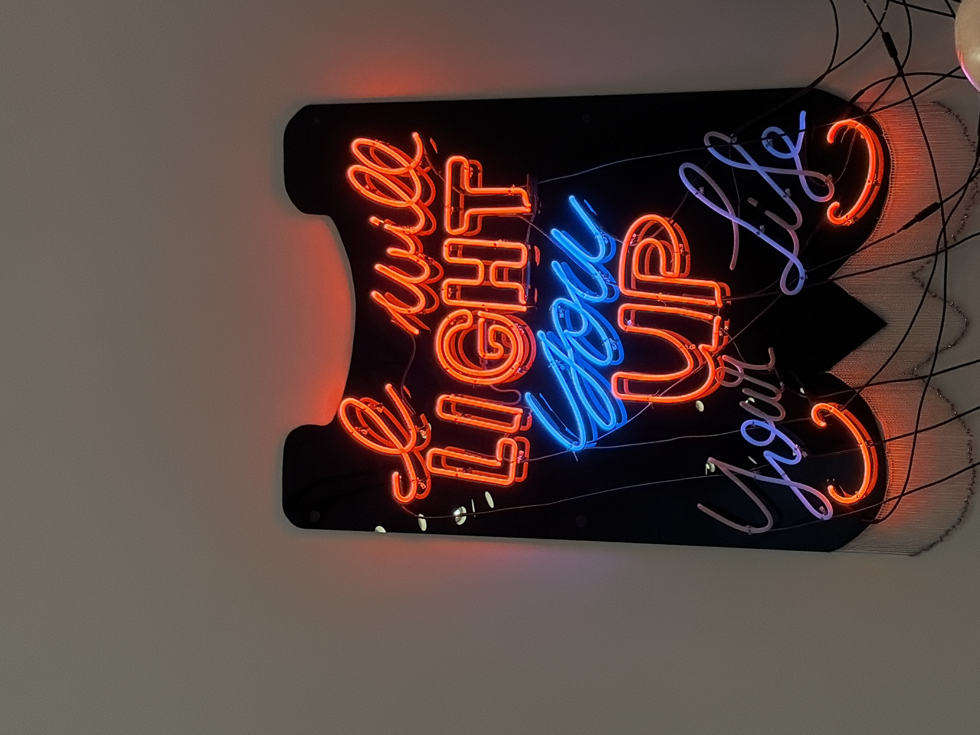 The image shows a neon sign mounted on a wall. The sign, in a black backboard, reads "I will LIGHT you UP your Life." The words "I will" and "LIGHT" are rendered in orange neon. "You Up" is in blue neon, and "Your Life" is again in orange. The sign appears to be illuminated. The wall is plain and light gray, with visible wiring and other decorative elements extending to the right side of the sign. The picture may have been taken indoors, and due to its style, may indicate a sign in Austin, Texas.