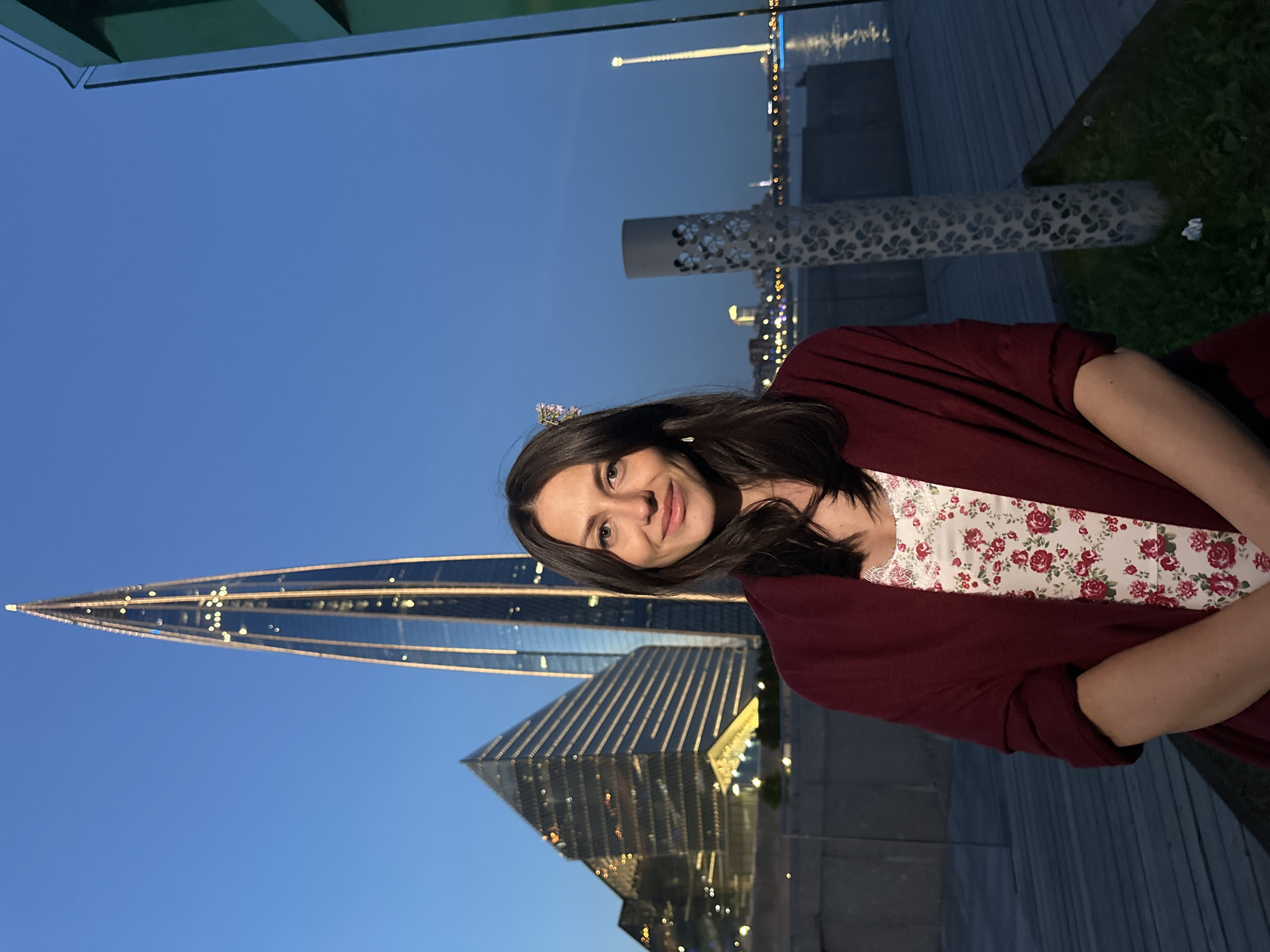 A medium shot captures a woman standing on a wooden deck at dusk, with a modern cityscape in the background. The woman, with dark hair and fair skin, is wearing a floral-print dress and a maroon shawl. She smiles at the camera. Behind her, a distinctive glass skyscraper with illuminated windows pierces the twilight sky, and a pyramid-shaped building with similar lighting is visible to its left. The scene appears to be the Lakhta Center area in Saint Petersburg, Russia, known for its contemporary architecture and waterfront views. The sky is a deep blue, transitioning to a darker hue, and the water in the distance reflects the city lights. A decorative cylindrical structure stands to the right of the woman, partially obscuring a tall, thin illuminated tower in the far background.