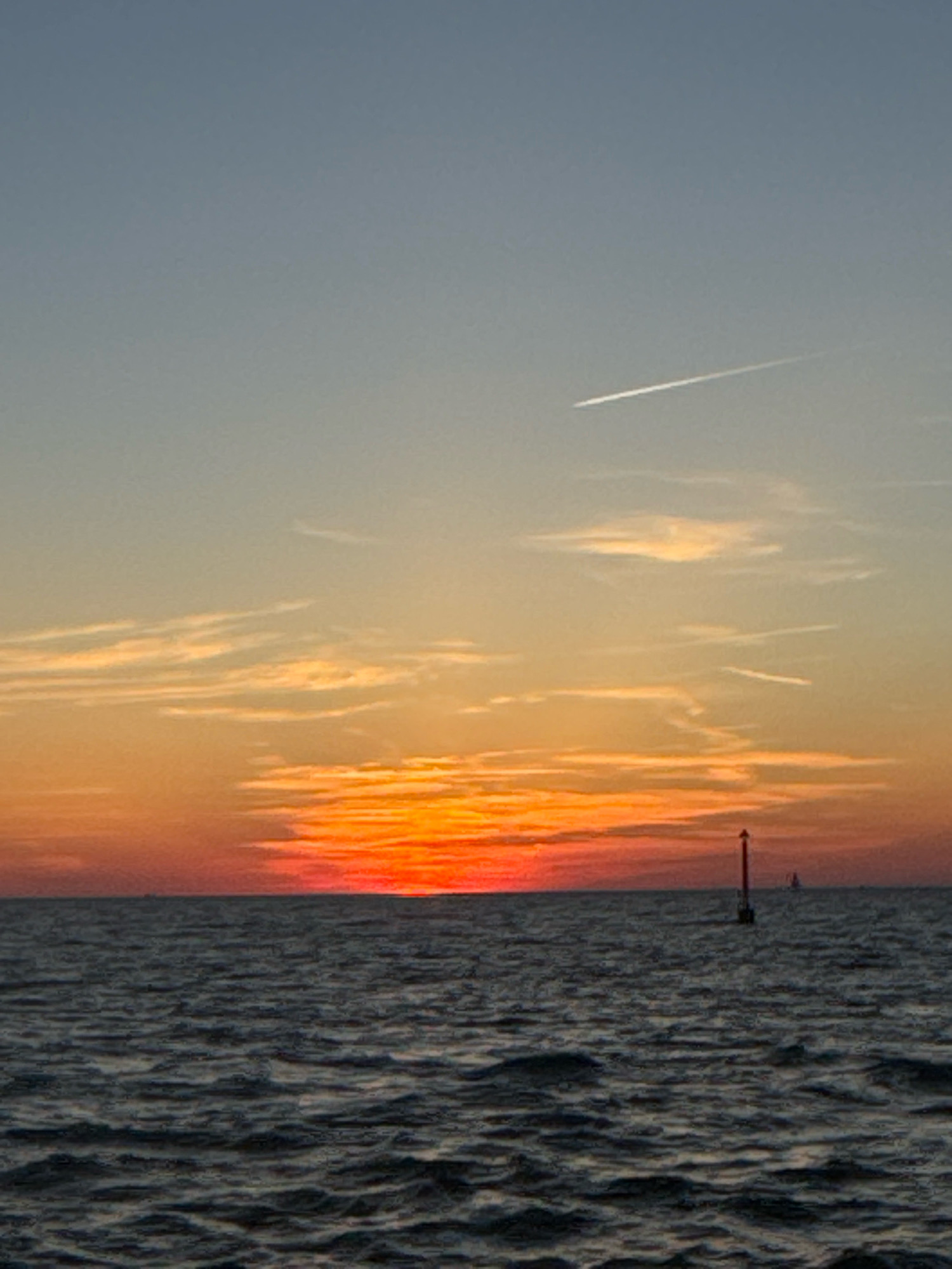 A wide-angle, vertically oriented, outdoor shot captures a sunset over a body of water. The sky is a gradient of light blue at the top, transitioning to yellow and then a vibrant orange and red near the horizon. Wispy clouds are scattered throughout the sky, and a faint airplane contrail is visible in the upper portion of the image.
The water is dark and choppy, with visible ripples and waves. In the middle ground, a red and black buoy stands tall in the water. In the distance, the faint silhouette of a sailboat can be seen. The overall scene suggests a peaceful, natural setting at dusk. The image does not contain any individuals or specific structures that would indicate a precise location within Novigrad, Croatia.