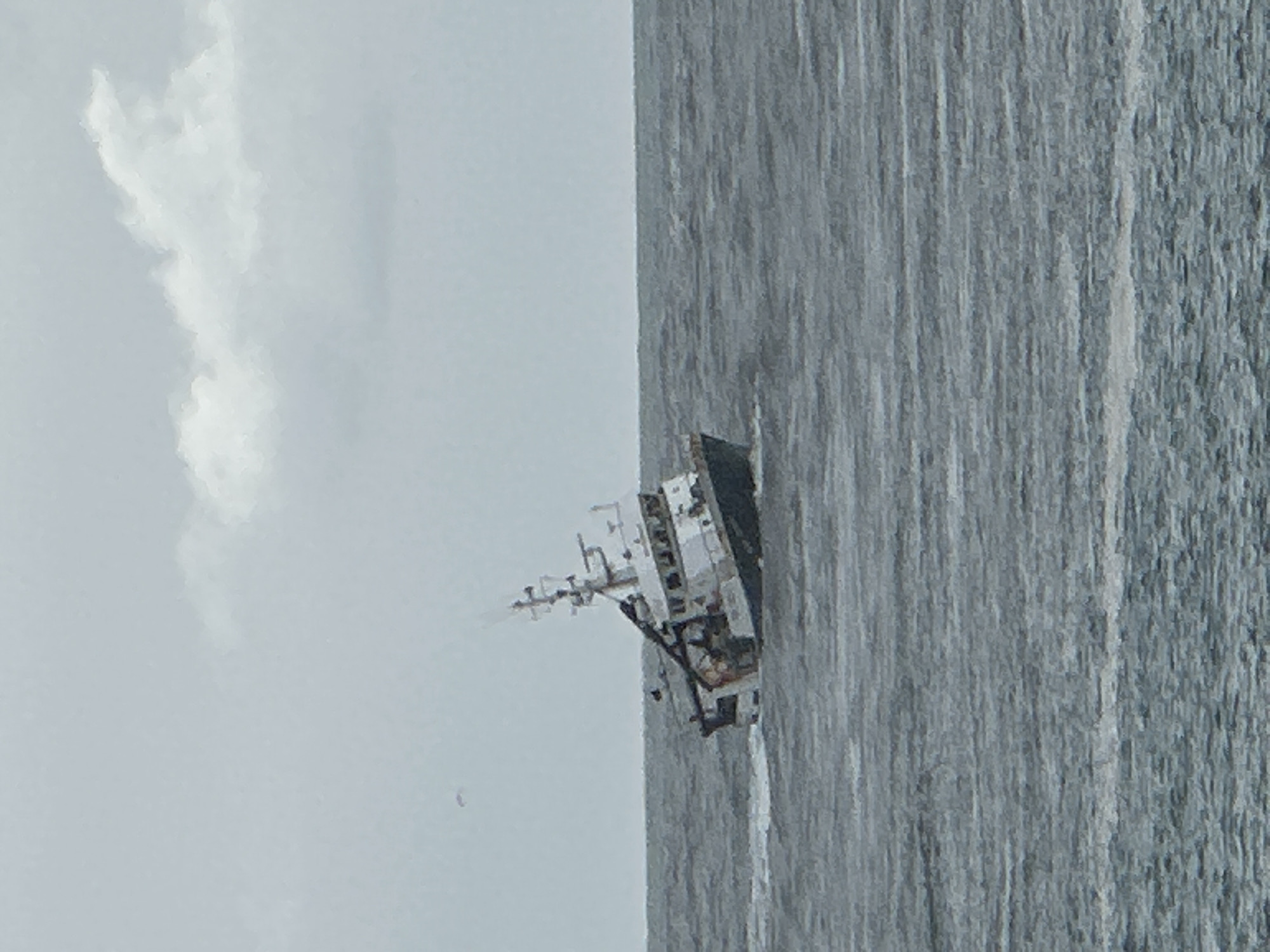 A commercial fishing vessel is seen on the sea off the coast of Gafanha da Nazaré, Portugal. The vessel is white with a tall mast. The sea is gray and choppy. The sky is overcast with a long cloud.