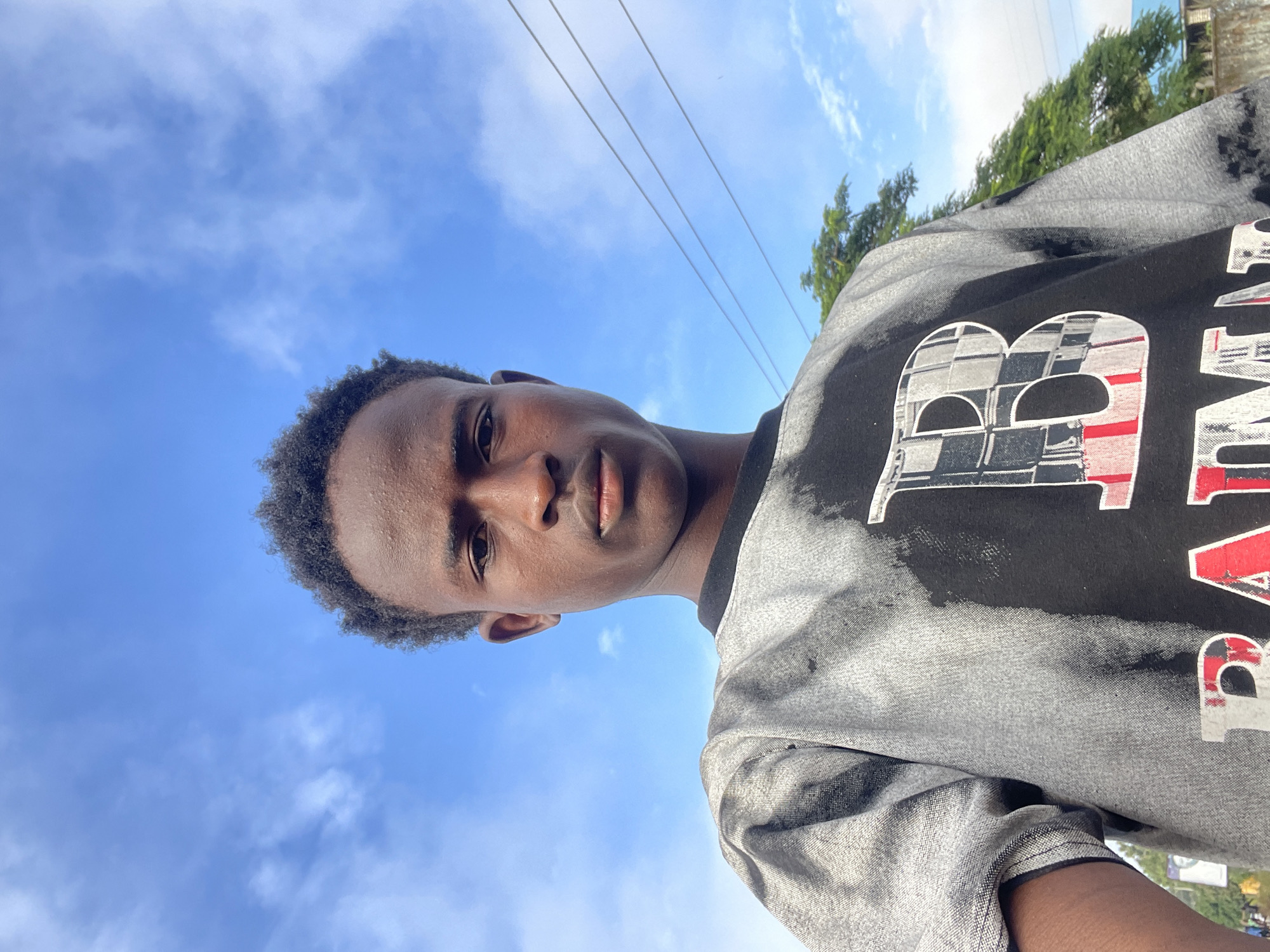 A young Black male is captured in a close-up, low-angle selfie, looking directly at the camera with a neutral expression. He has dark, curly hair and dark eyes. He is wearing a black t-shirt with a large, white, checkered 'B' printed on it, outlined in red. The t-shirt has a distressed, tie-dye-like pattern in shades of grey and black. The background is a bright blue sky with scattered white clouds. Three parallel utility wires are visible diagonally across the upper right quadrant of the image. To the right, a lush green tree with dense foliage is partially visible, and beyond it, a glimpse of a building and a hint of a blue roof can be seen. The overall impression is an outdoor setting, possibly in an urban environment within Jos, Nigeria, given the presence of utility wires and building structures.