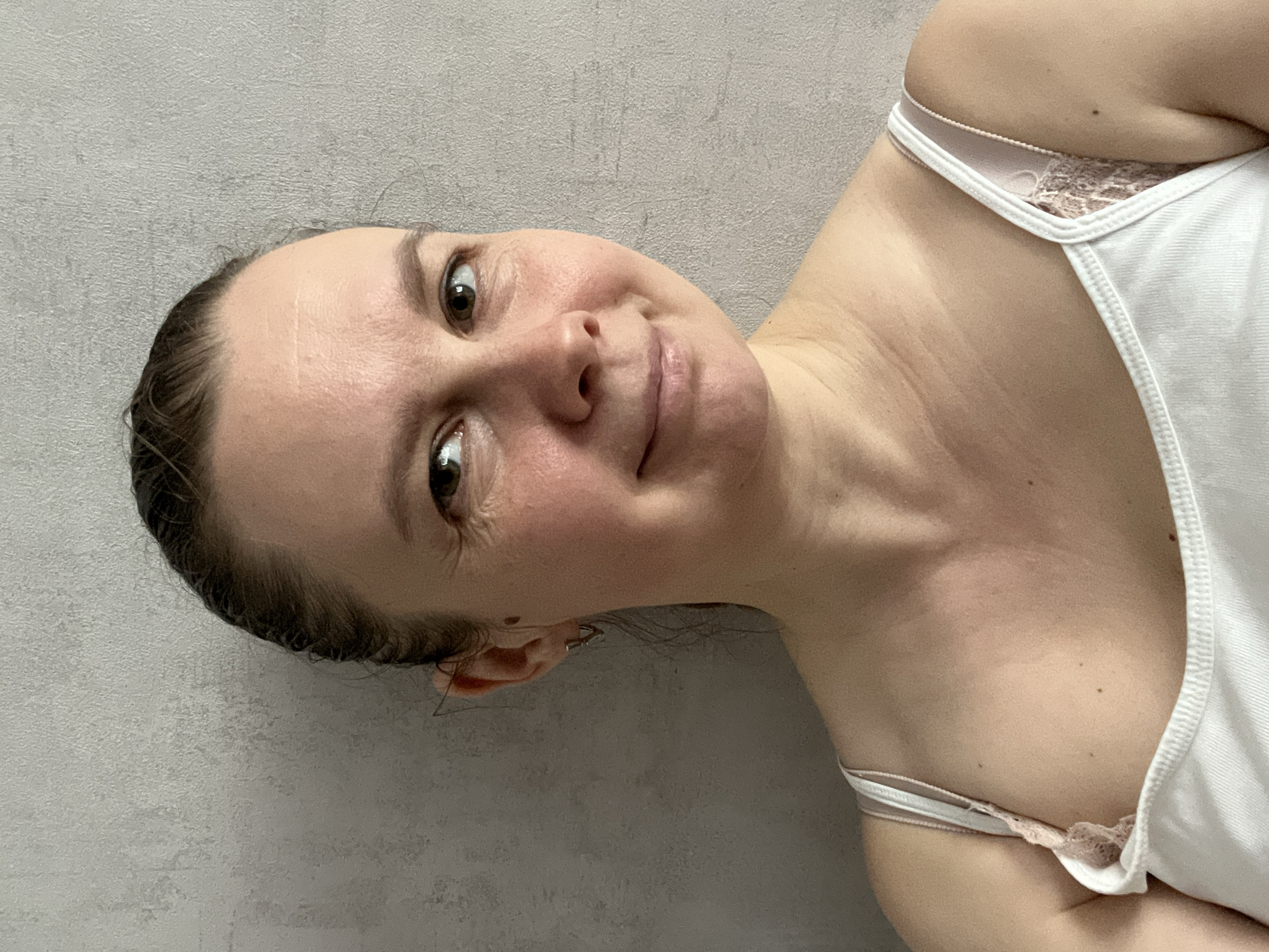 A woman is posing for a selfie against a gray wall. She is wearing a white tank top with lace trim. Her hair is pulled back, and she is wearing small earrings. The location in the city of Izhevsk, Russia is not discernible from the image.