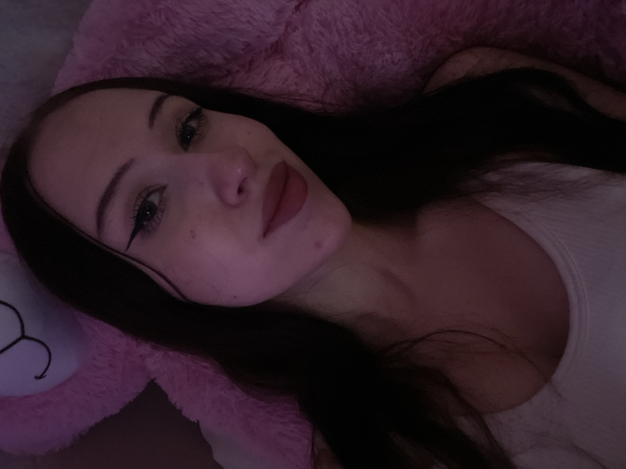 The image is a close-up, overhead shot of a young woman with dark hair and fair skin. She is wearing a white tank top. She has makeup on, including eyeliner and lipstick. She is lying down, leaning against a large pink stuffed animal, possibly a bear, which is partially visible in the background. The lighting is dim and appears to be coming from a pink-toned source, casting a soft glow on the scene. The image does not provide any information about the location being in Votkinsk, Russia.