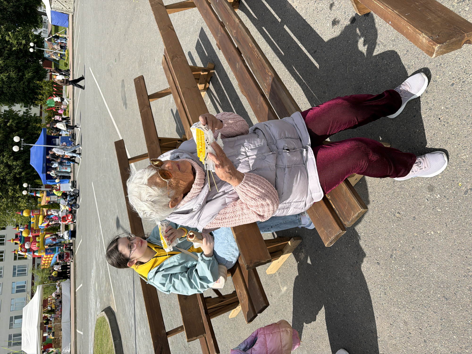 A high-angle shot shows two individuals seated on a wooden bench. One, possibly an elderly person, wears an old-age makeup prosthetic and costume, with a white wig, large sunglasses, and a white puffy vest over a pink sweater, maroon pants, and white sneakers. The second person, likely a younger individual, is seated to the left of the first. They are wearing eyeglasses, a yellow shirt, and a light blue jacket. Both individuals are holding food, which appears to be corn on the cob, in their hands.
The wooden bench consists of multiple planks of wood arranged at varying heights. The scene is situated in an open, paved area, likely outdoors, with other structures and people visible in the background. In the distance, there is a playground with an inflatable structure, several tents, and a number of people walking around. A multi-story building is visible on the left-hand side of the frame.
Based on the available information, the location is identified as Valozhyn, Belarus.