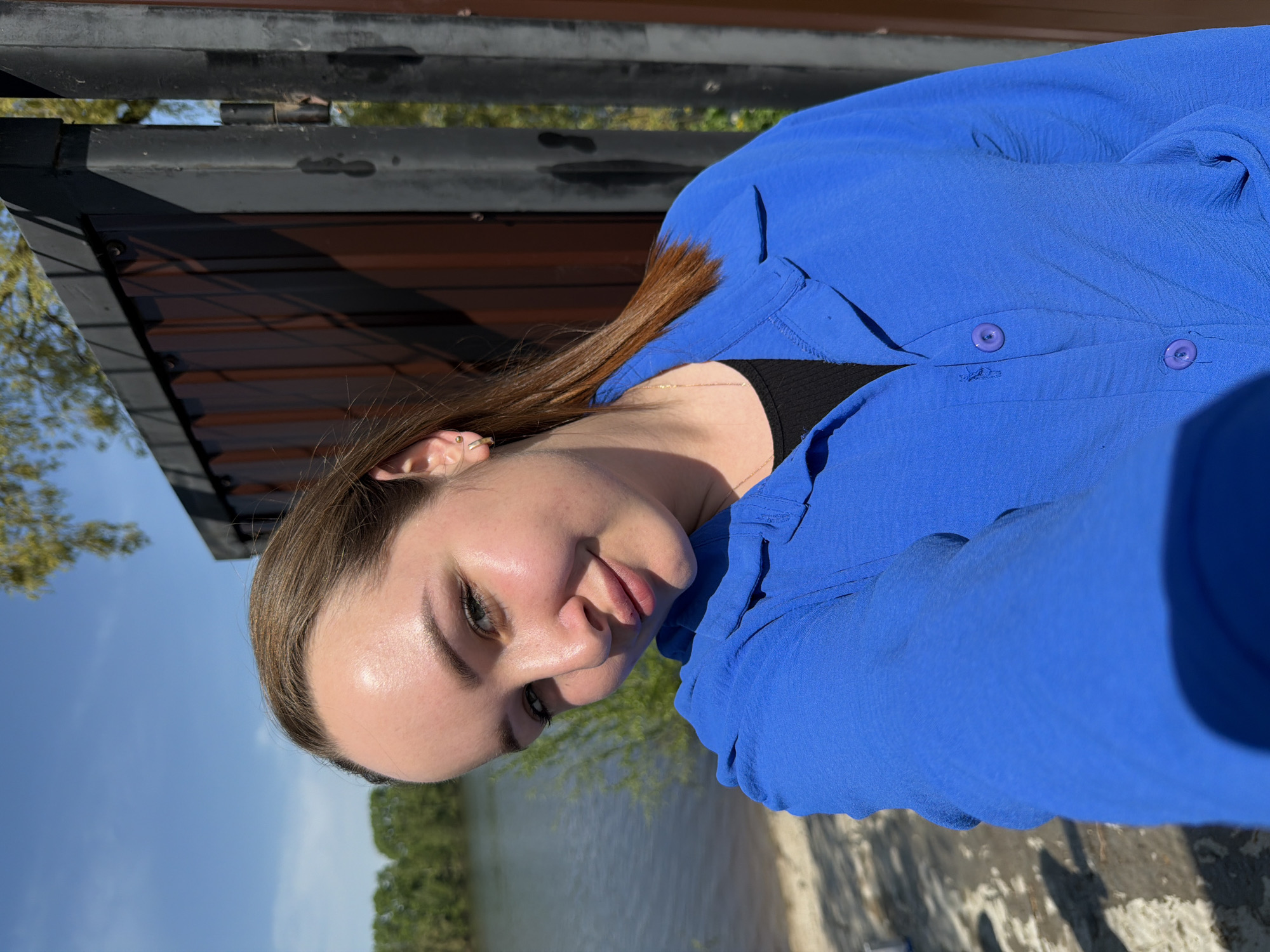 A young woman with light skin and brown hair is sitting outside, likely by a body of water. She wears a blue button-down shirt. The background shows what appears to be a lake or wide river, and a wooded area in the distance under a clear, sunny sky. Some architectural structures, possibly related to a park or building, are also visible in the background. It is not possible to confirm the location in the city of Chaikovsky, Russia.